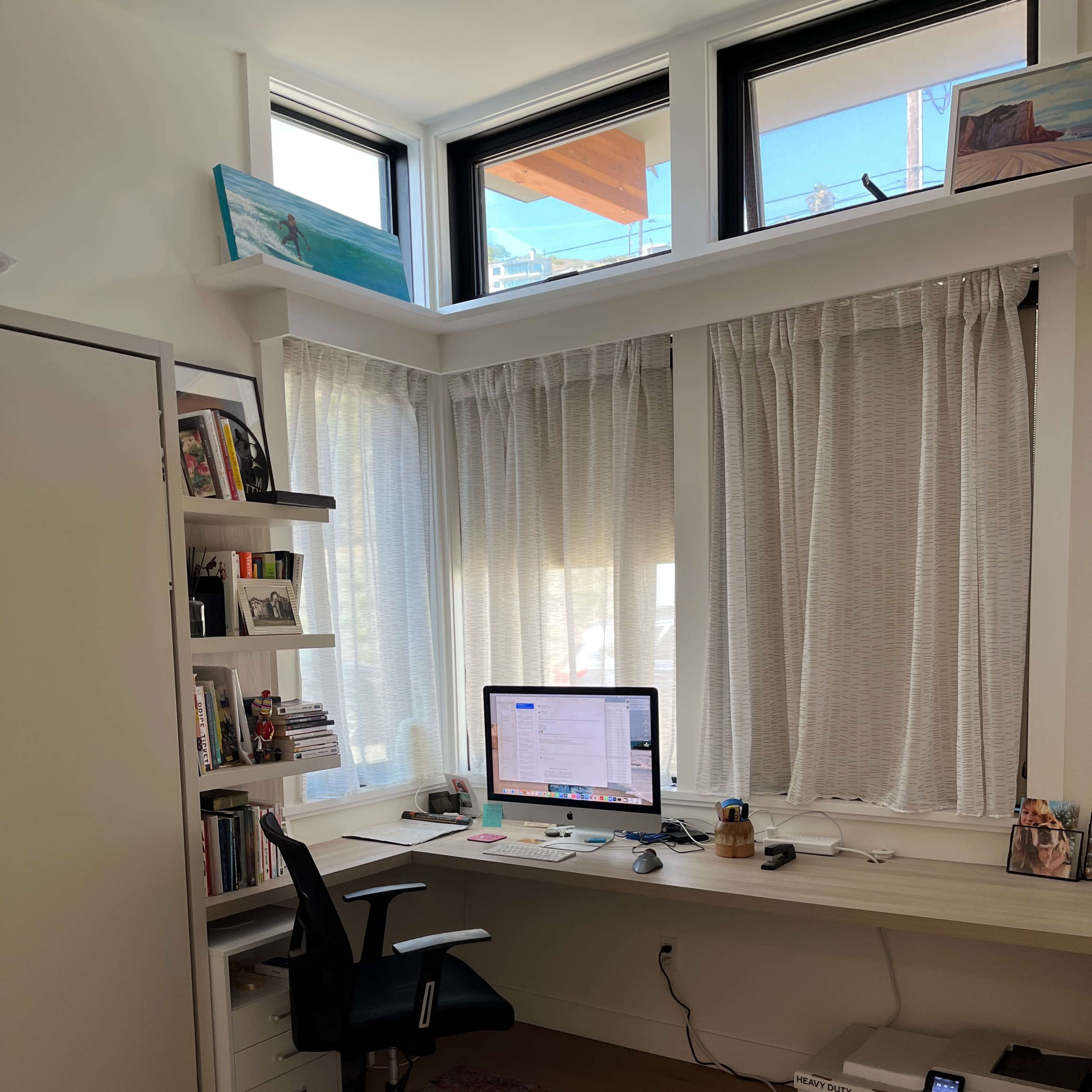 A tidy home office features a desk with a computer, a rolling chair, and bookshelves, situated near large windows with sheer curtains.