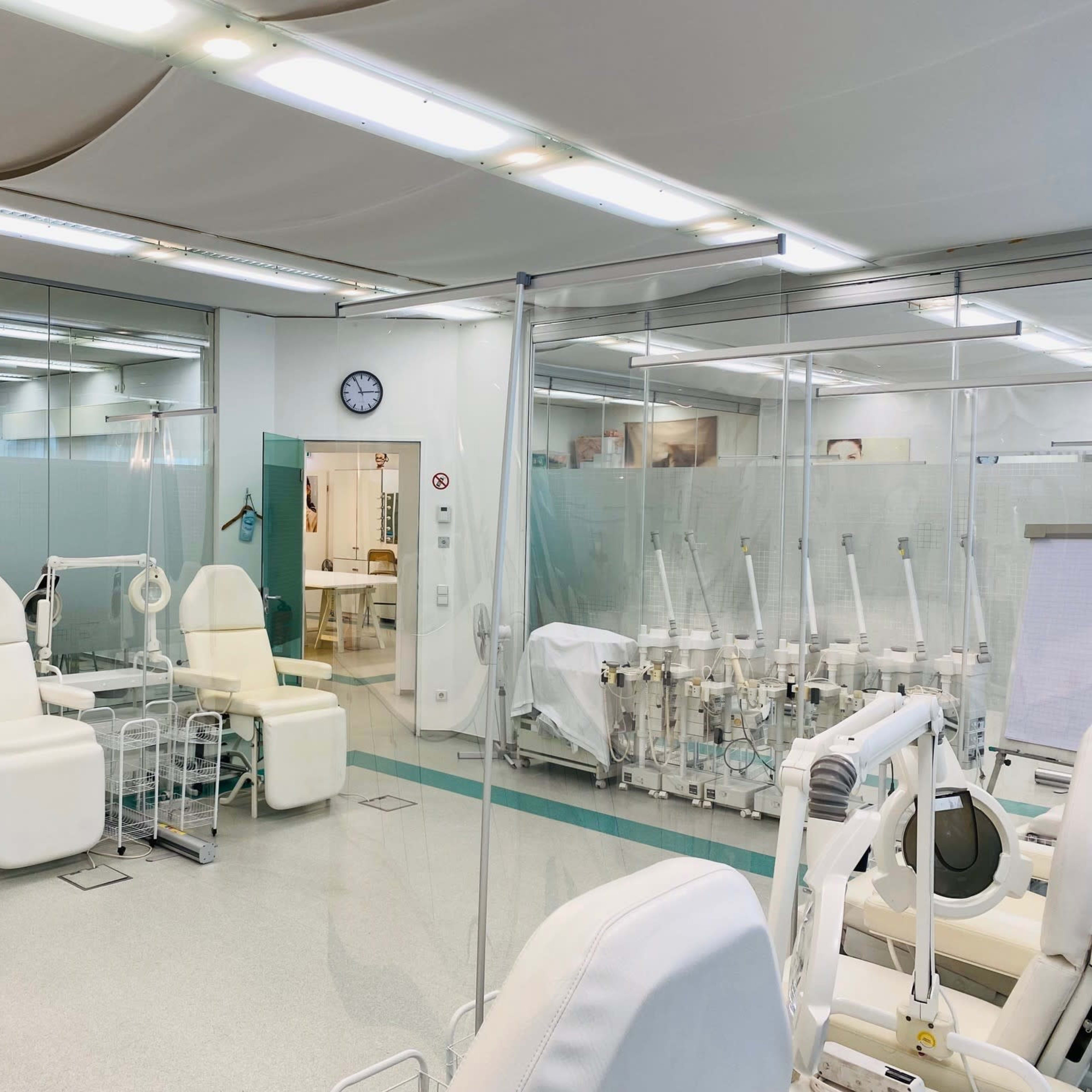 A medical room with multiple treatment stations separated by clear partitions, featuring white reclining chairs and medical equipment in the background.