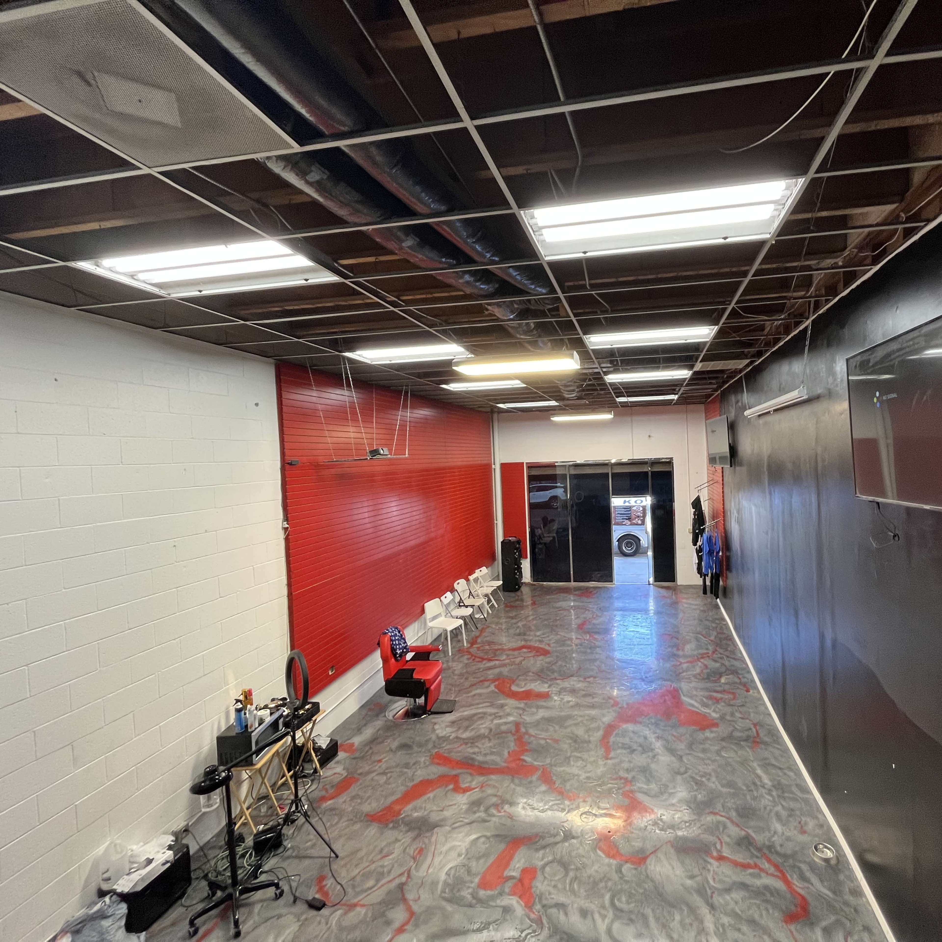 An interior of a room with a red accent wall, a polished floor, and several chairs along one side, with exposed ceiling beams and overhead lighting.