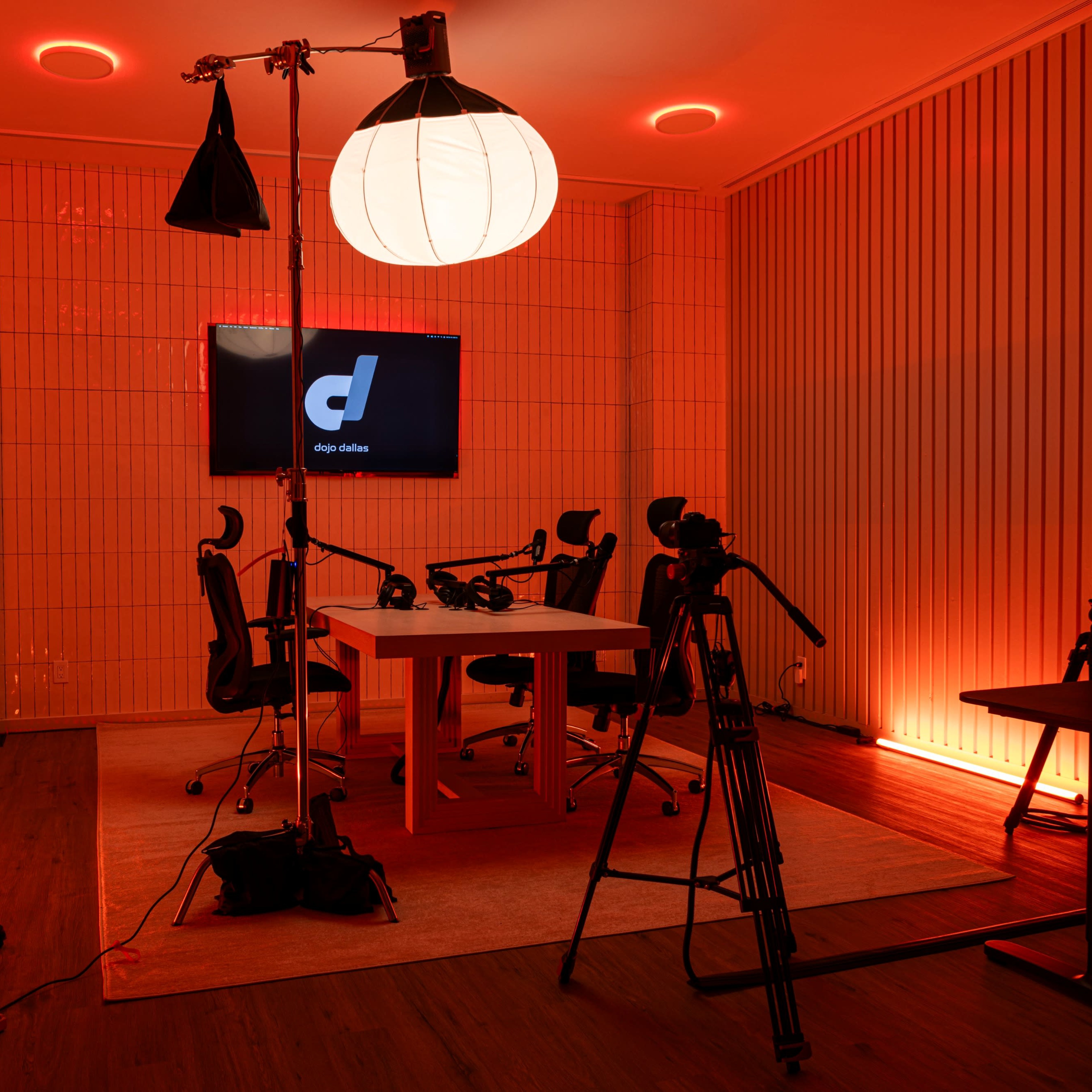 A modern recording studio with a large table, chairs, multiple cameras, and a bright circular light, all set against an orange backdrop.