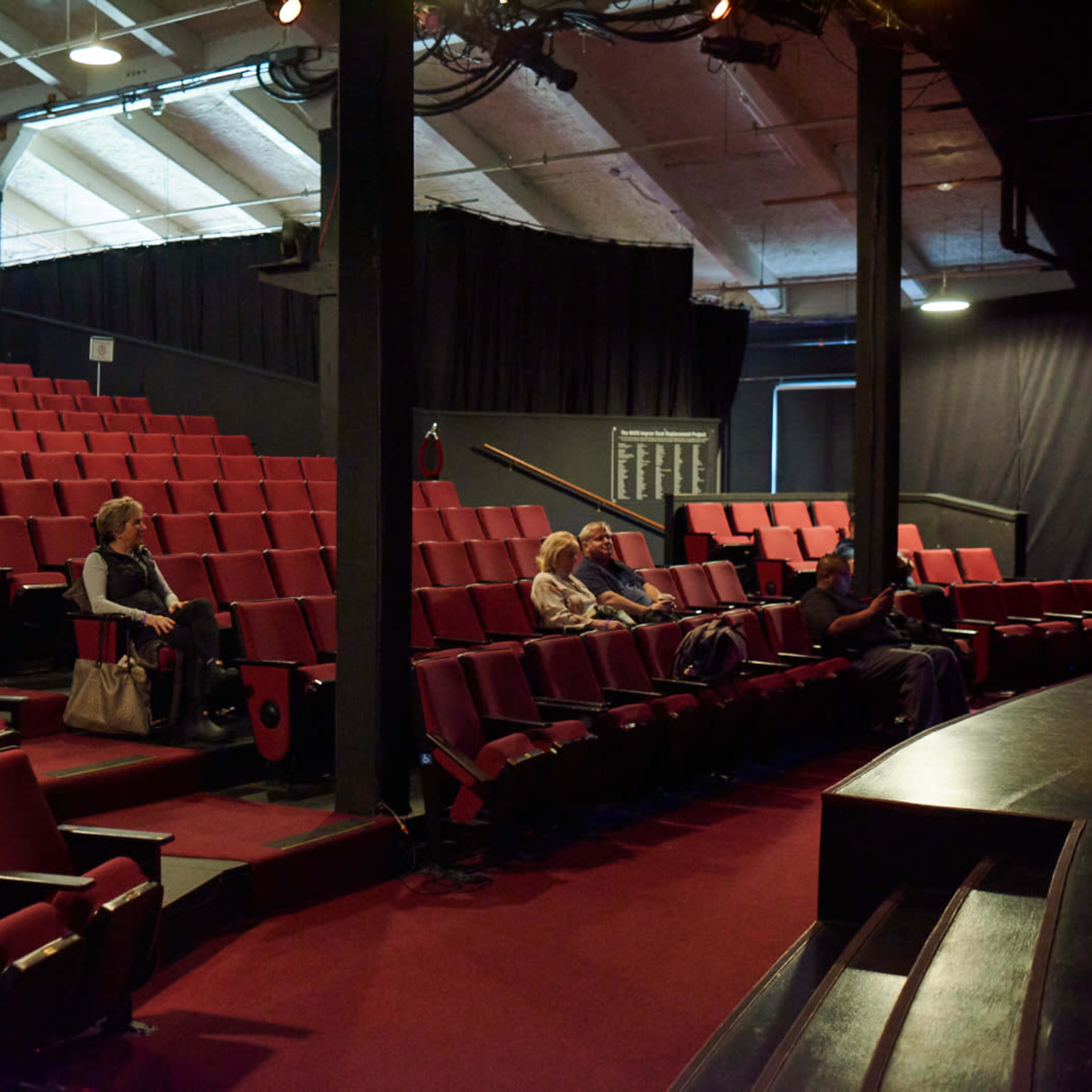 An empty theater with red seats and a sparse audience seated in the front rows.