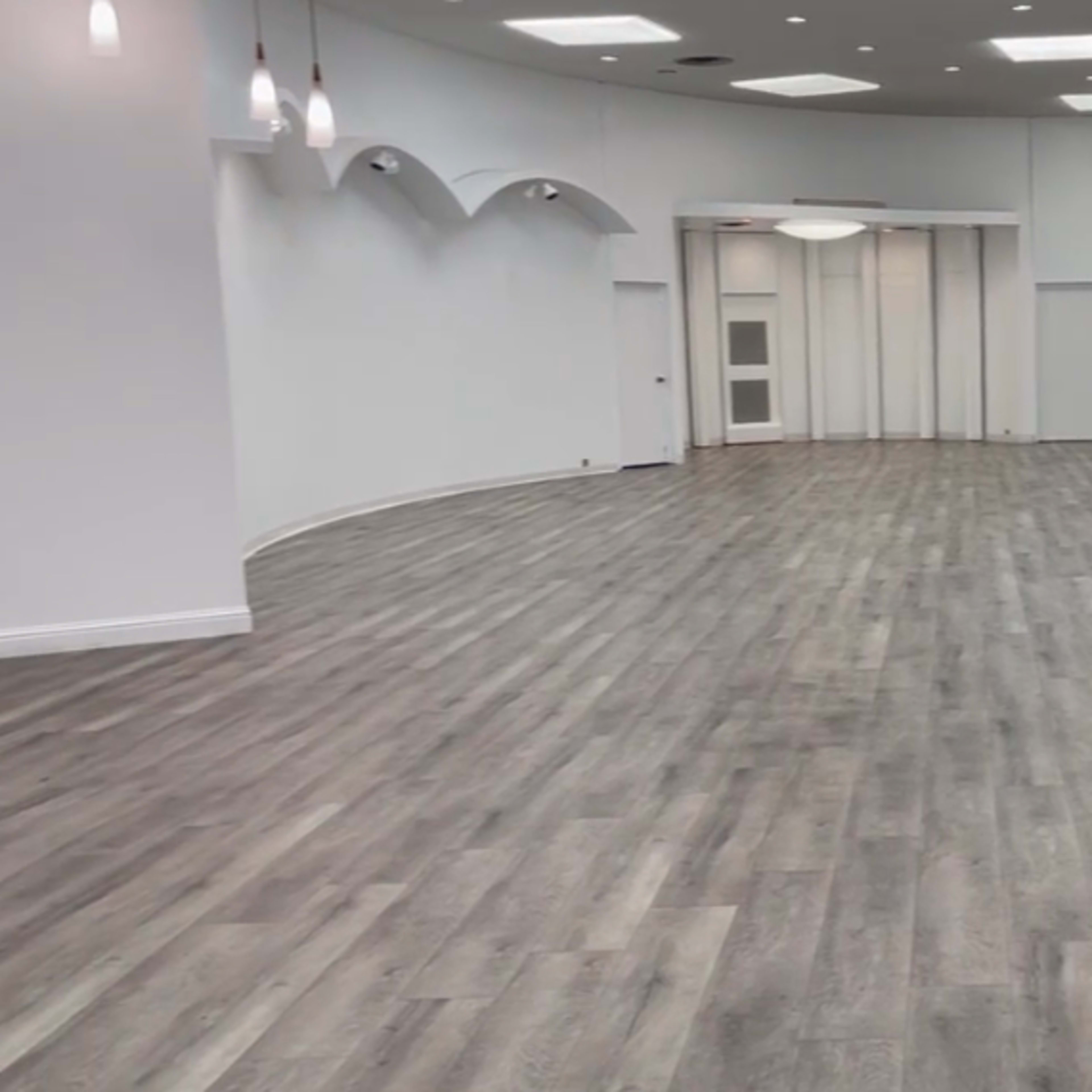 A spacious, empty room with light-colored wooden flooring and white walls, featuring multiple doorways and overhead lighting.