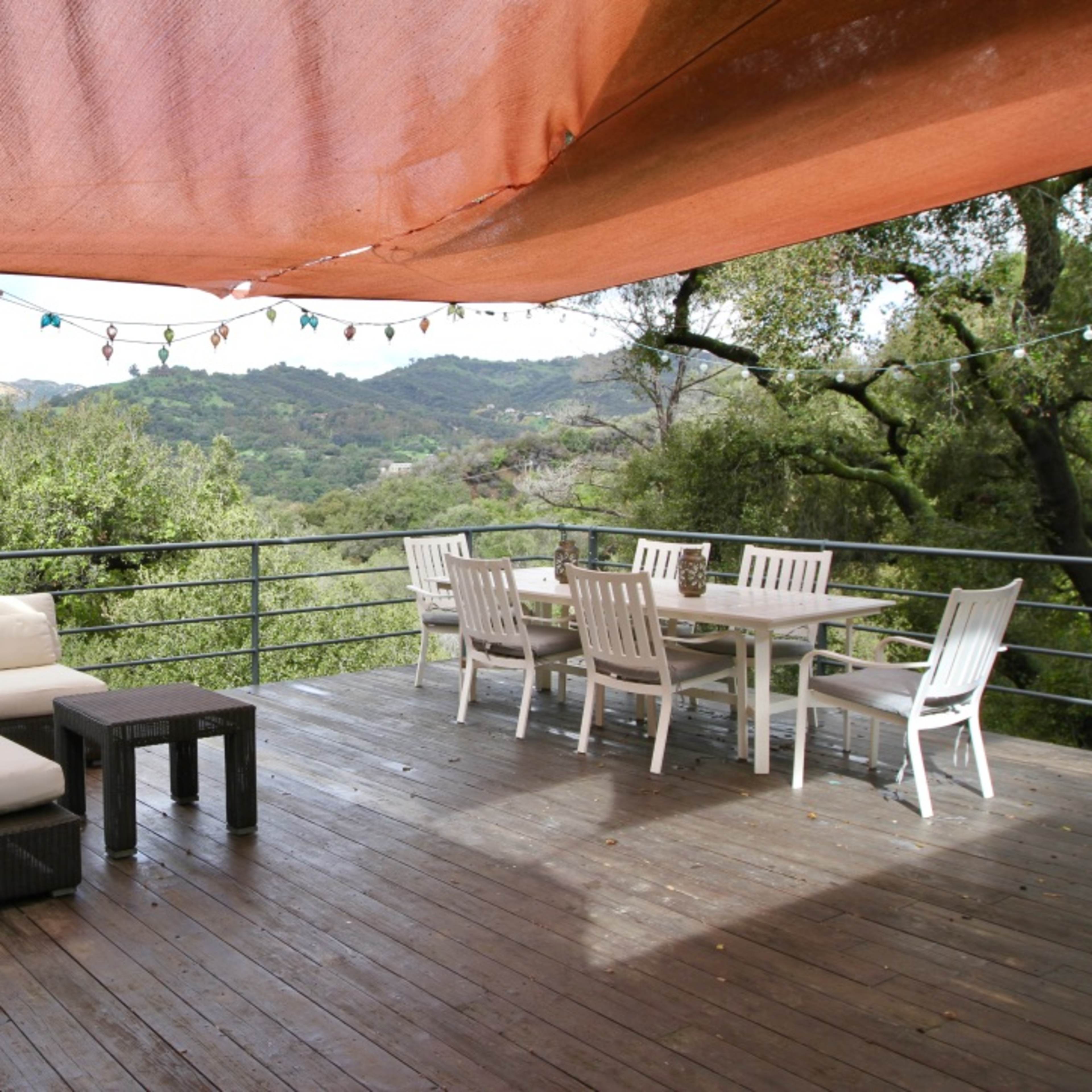 A wooden deck features a seating area with a couch and table, along with a dining set, all set against a backdrop of lush greenery and rolling hills.