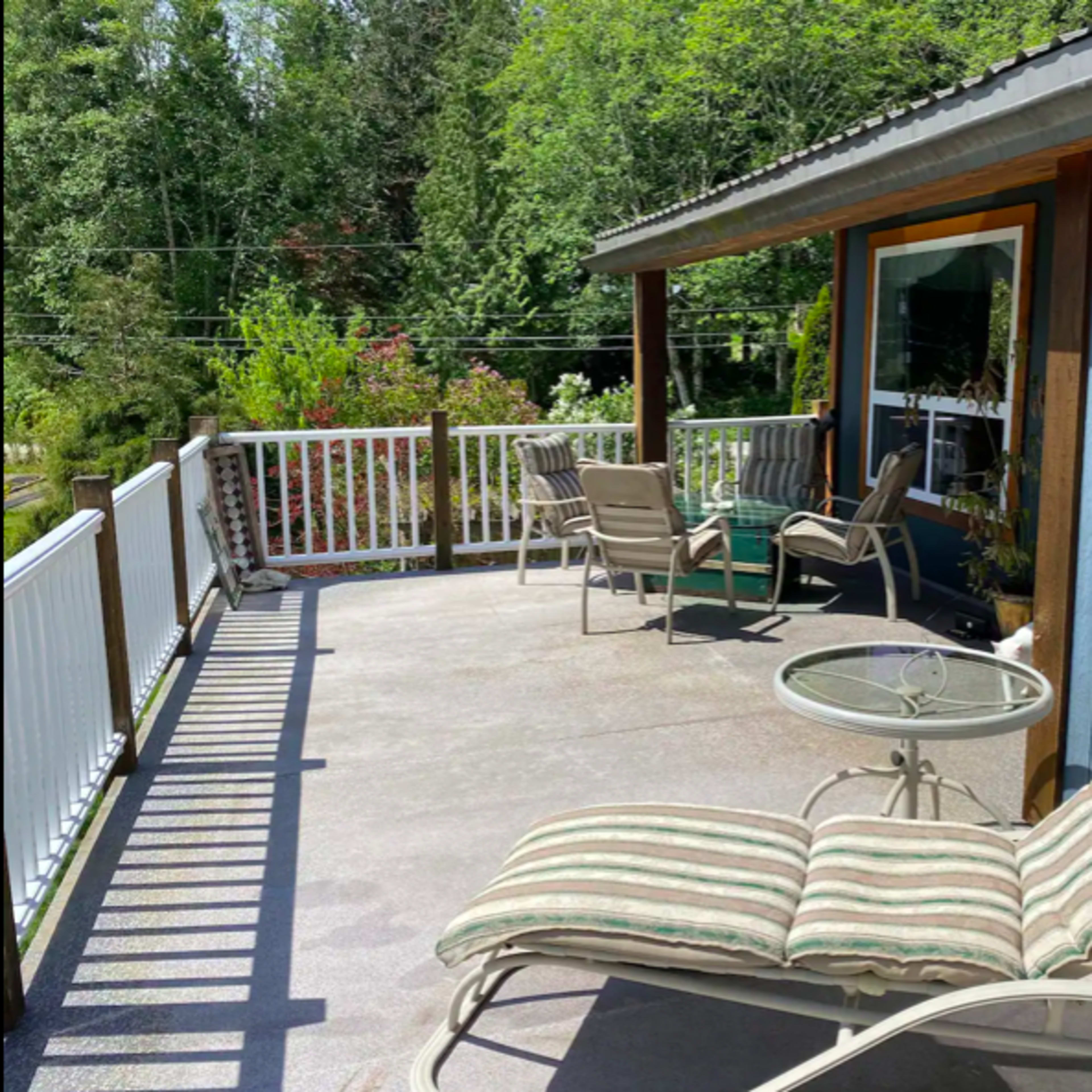 A sunlit deck features a lounge chair, a small table, and several seating options by a house surrounded by greenery.