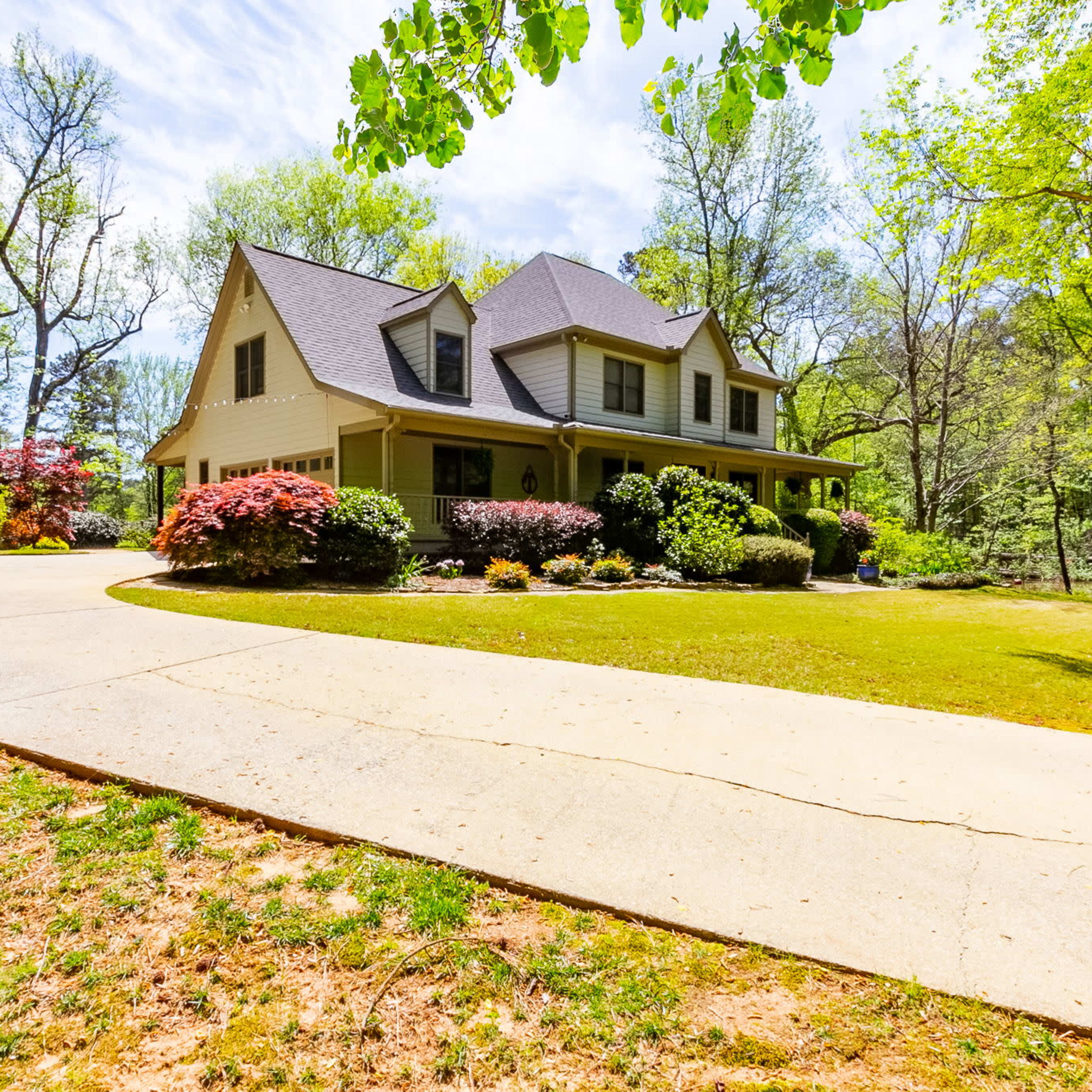A landscaped driveway leads to a two-story house surrounded by trees and flowering shrubs.