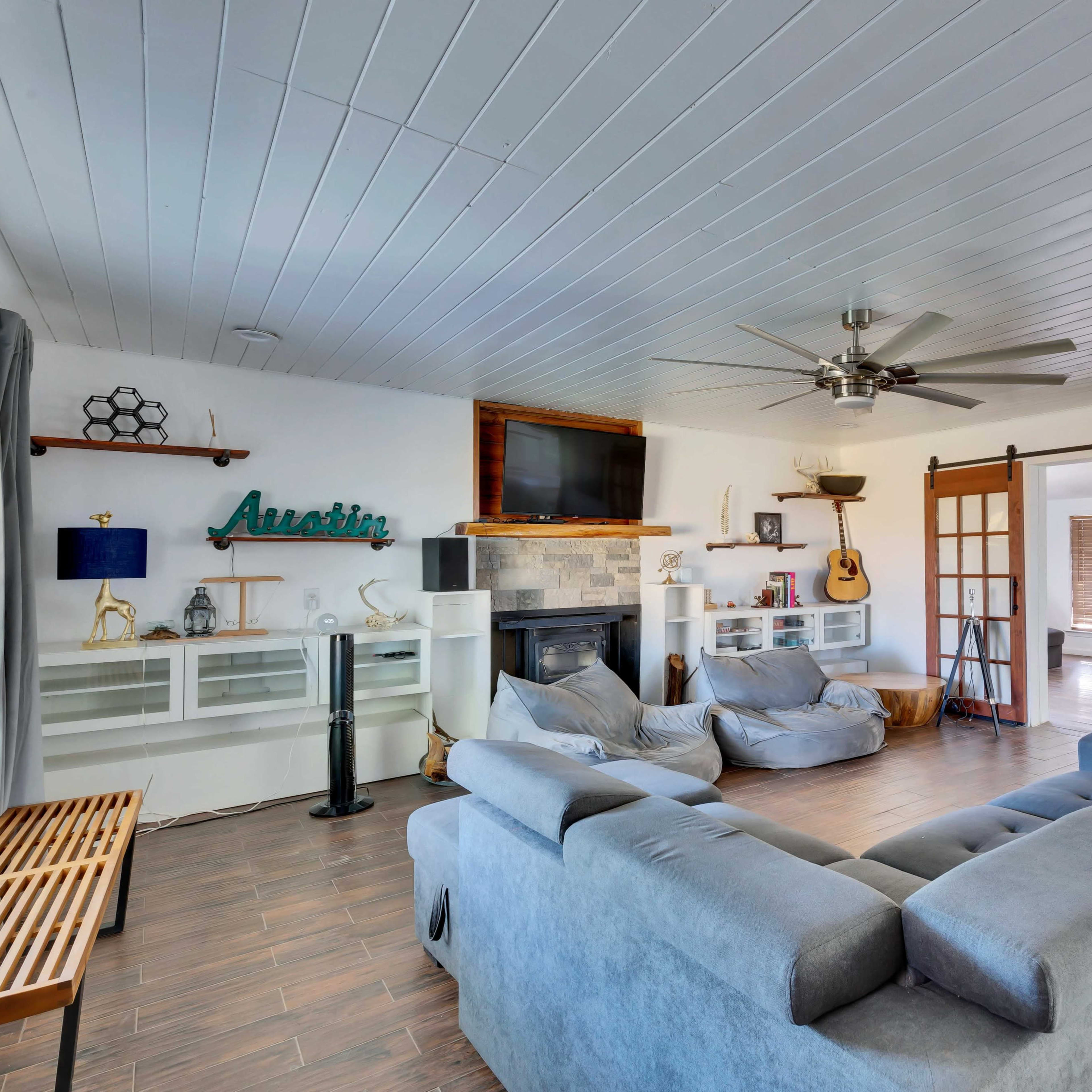 A spacious living room features a ceiling fan, a gray sectional sofa, and a fireplace, with large windows and a set of sliding doors leading to another area.