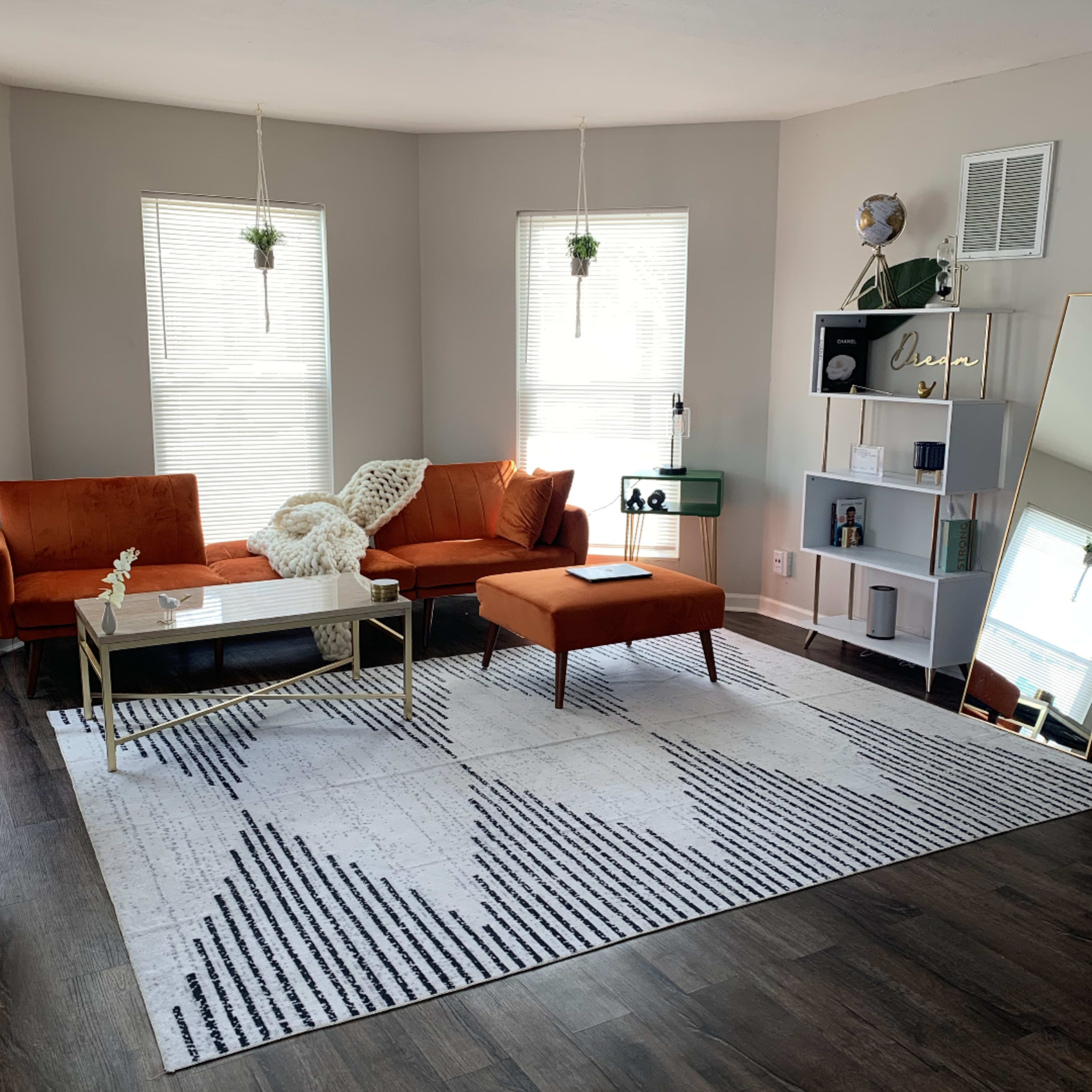 The living room features a pair of orange sofas, a coffee table, a patterned rug, and a bookshelf against the wall, illuminated by natural light from two windows.