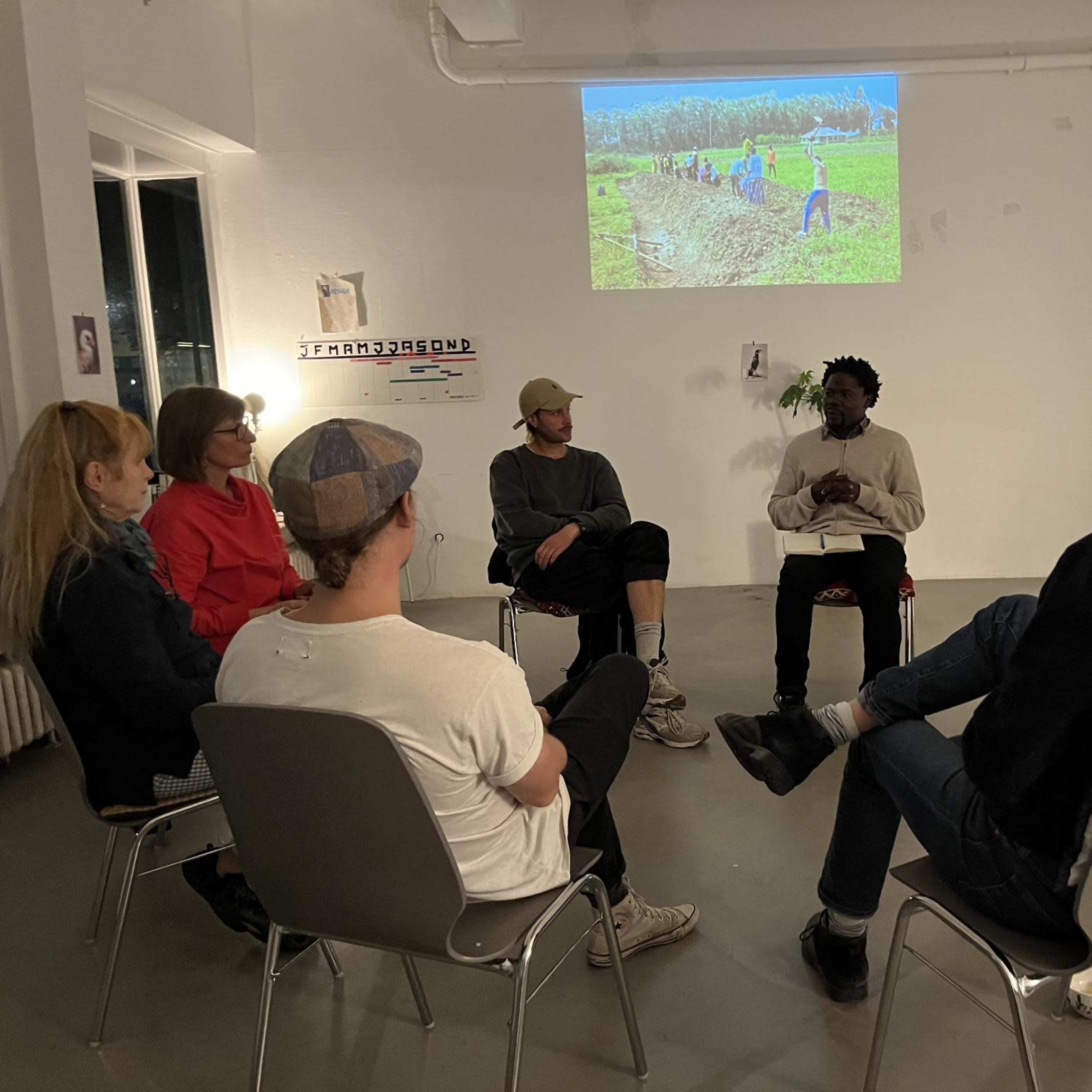 A group of people sits in a circle in an art space, listening to a speaker while a video plays on the wall behind them.