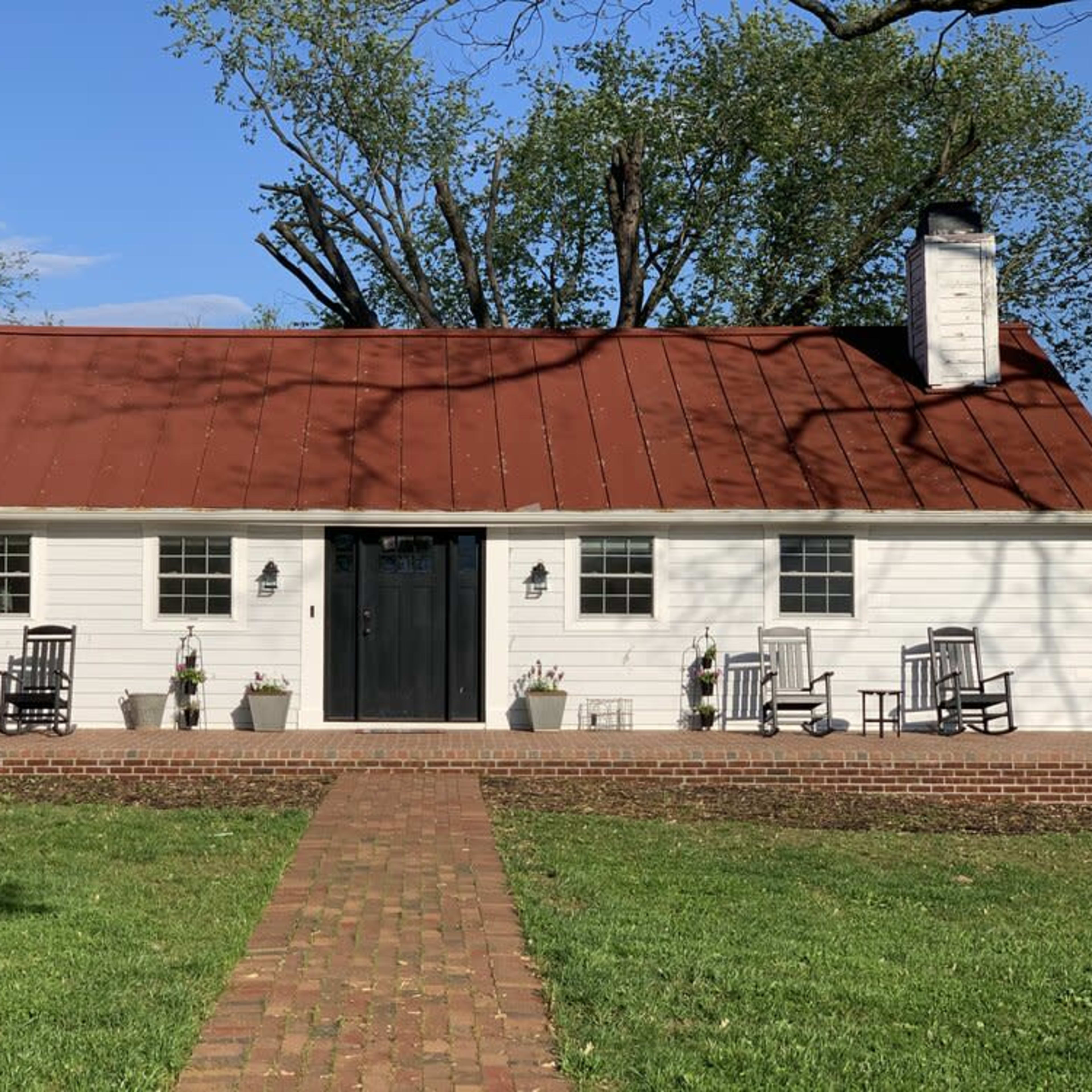 A white house with a red metal roof, a black front door, and a brick pathway leading to a covered porch with rocking chairs and potted plants.