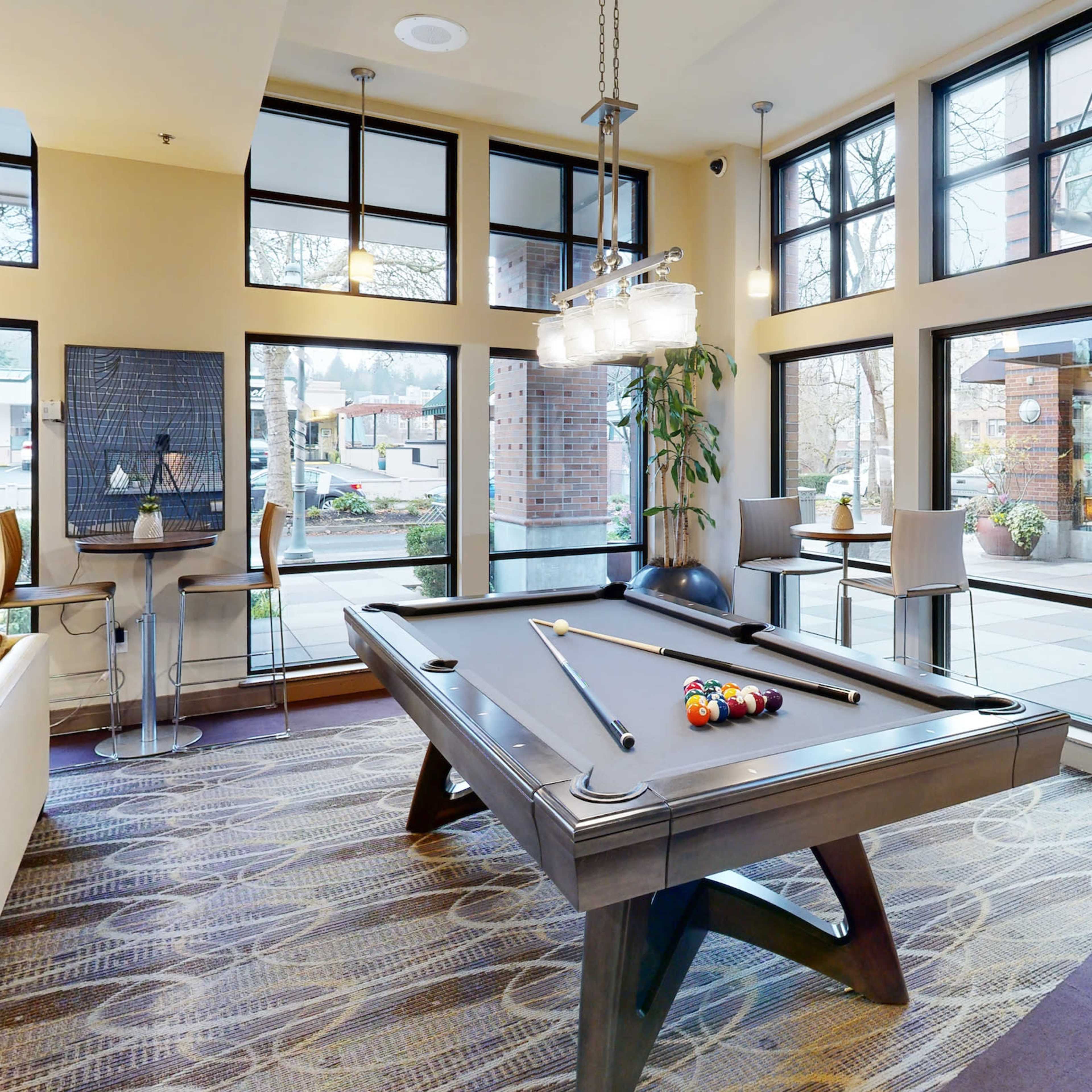 A well-lit lounge area featuring a billiards table, surrounded by large windows and modern furniture.