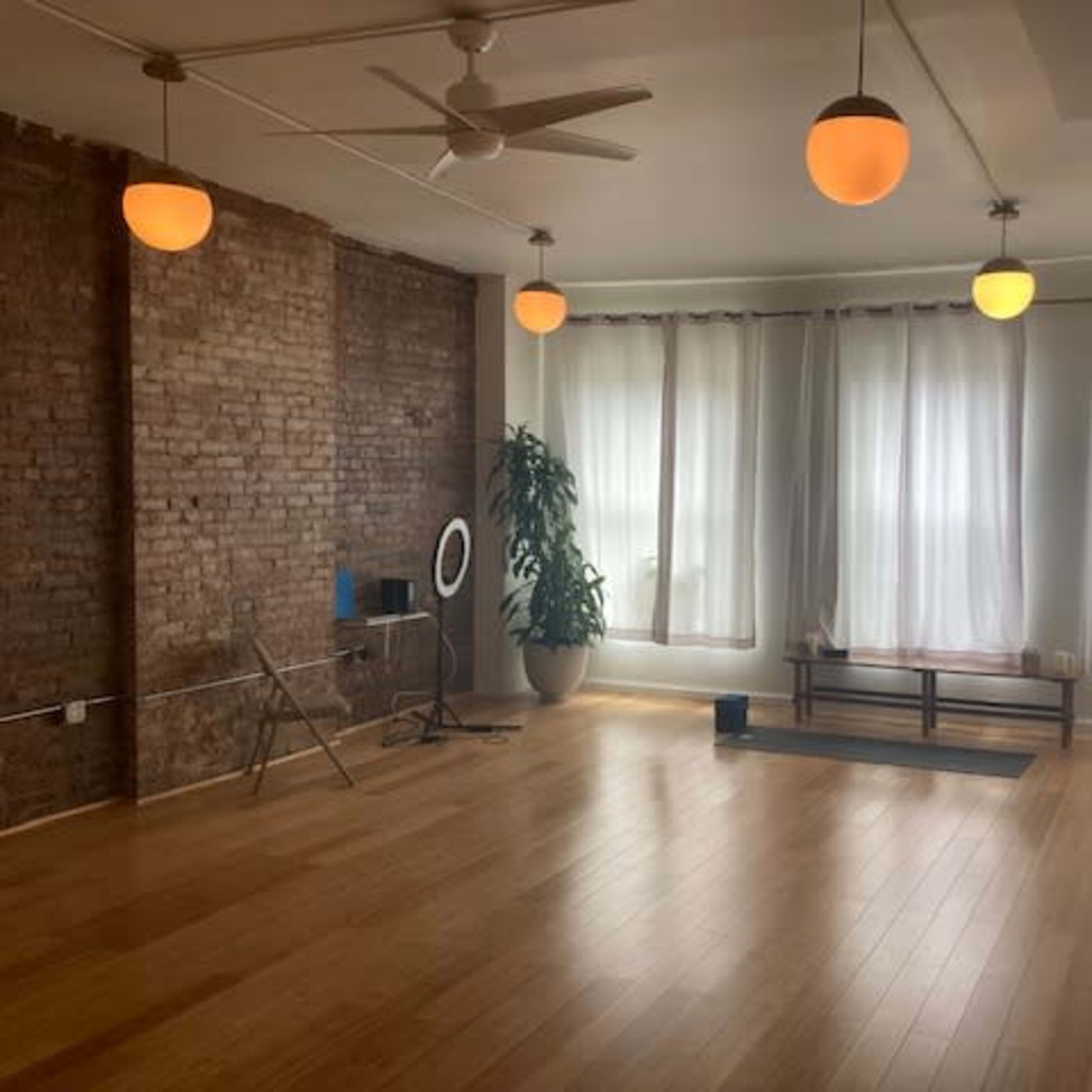 A bright, spacious room with wooden floors, large windows covered by sheer white curtains, and warm pendant lights hanging from the ceiling.