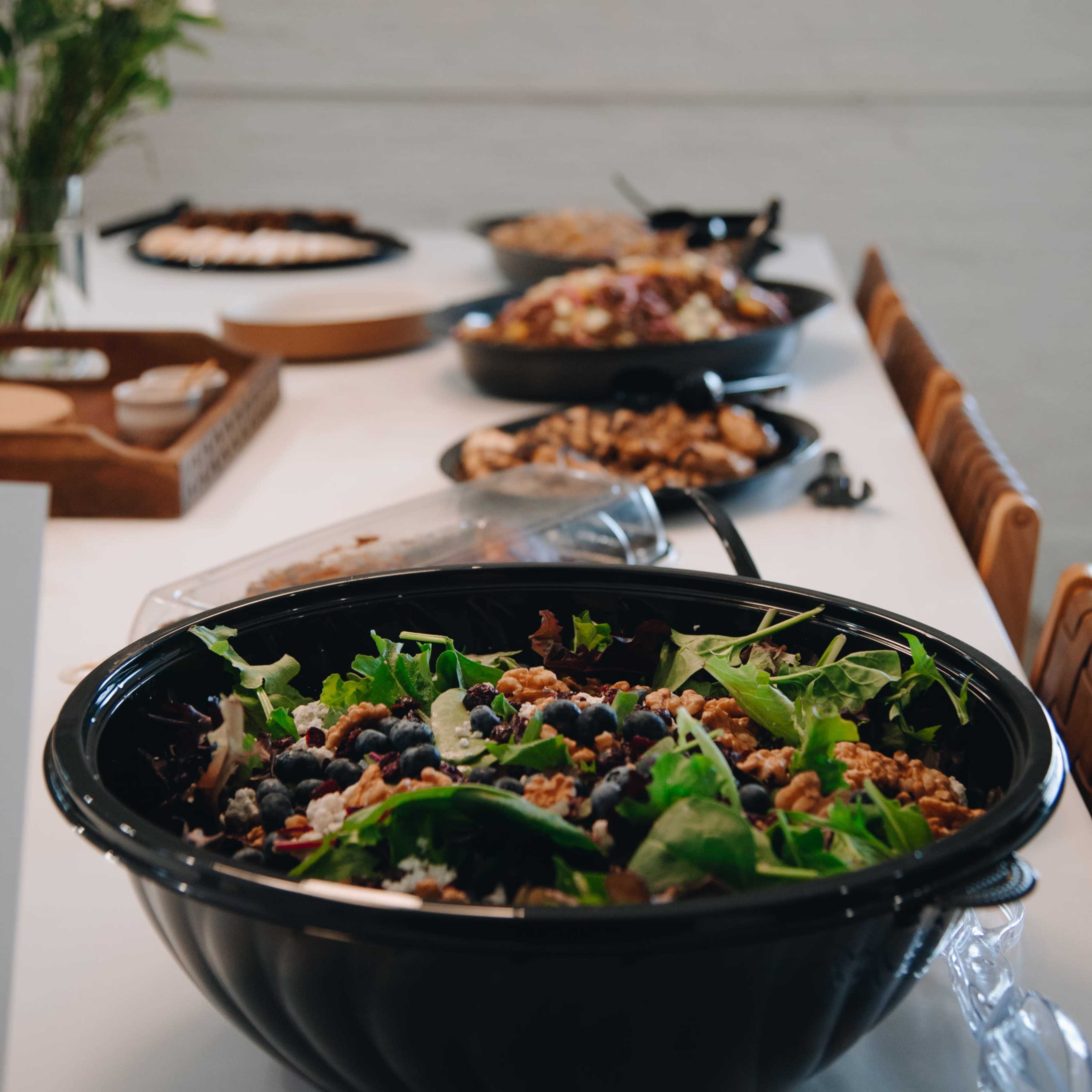 A large, black bowl of salad sits in the foreground with various dishes arranged on a table in the background.