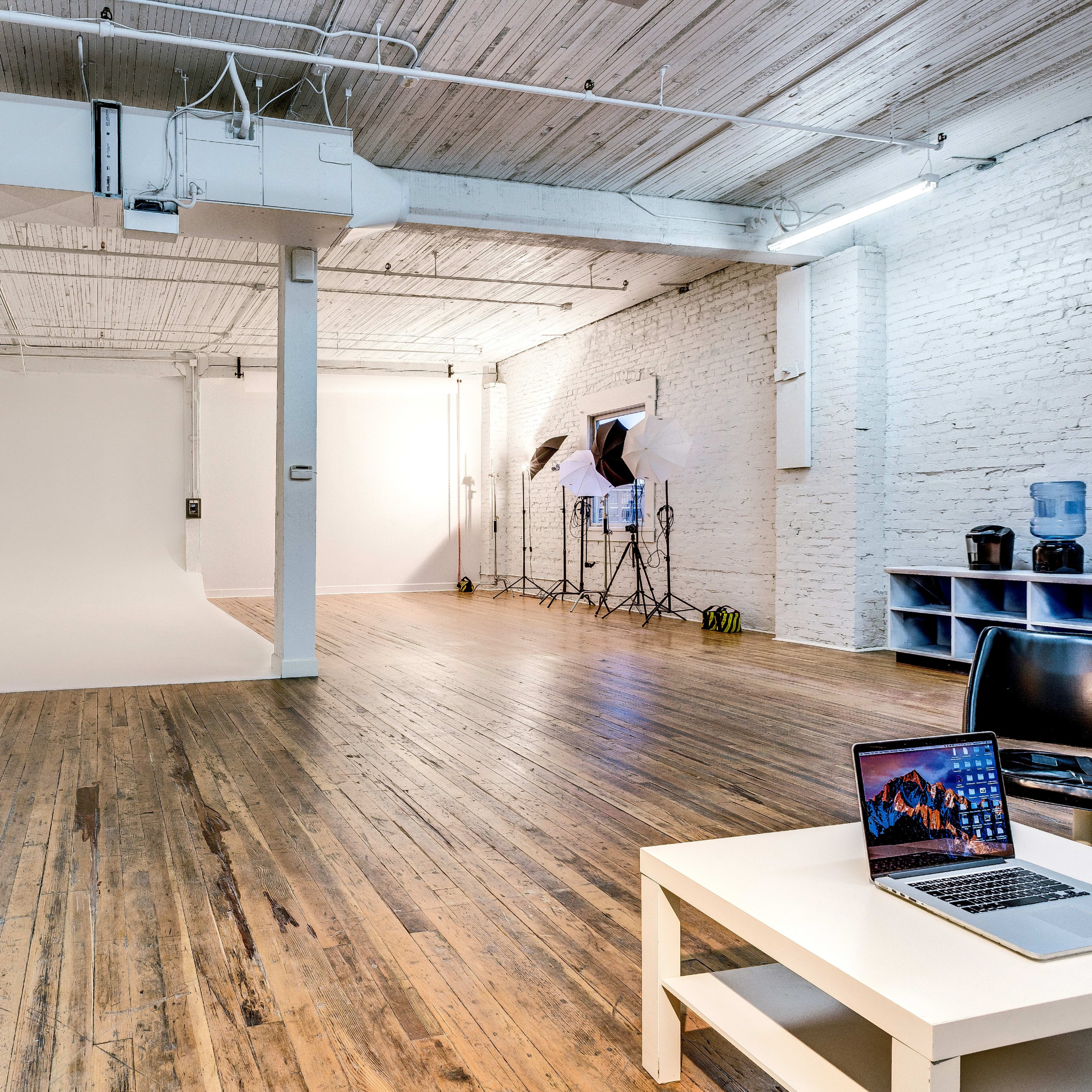 A spacious studio with wooden floors, white brick walls, and a large blank backdrop, accompanied by photography equipment and a workspace with a laptop.