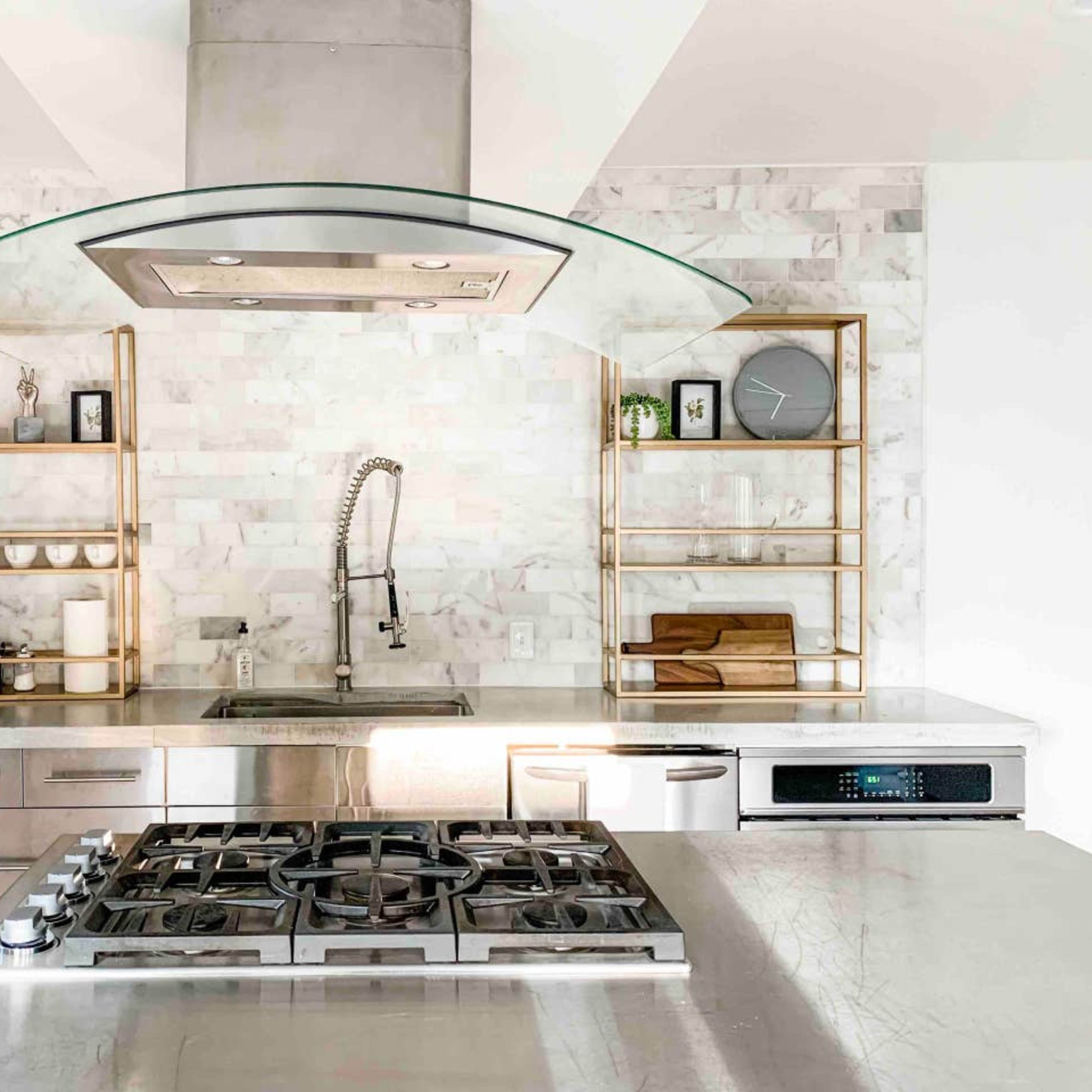A modern kitchen featuring stainless steel appliances, a gas stovetop, a marble backsplash, and open metal shelving displaying dishware and utensils.