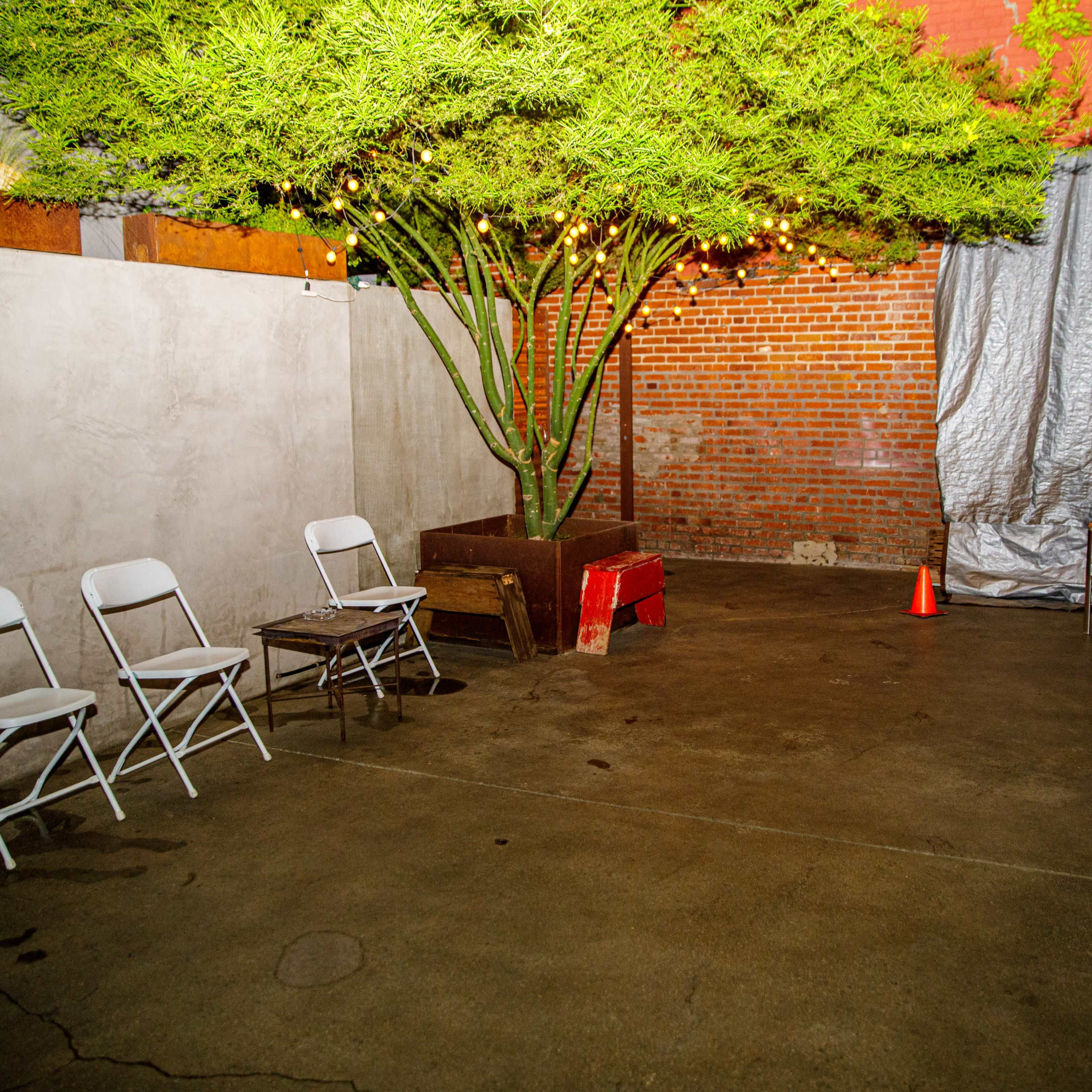 An empty courtyard with folding chairs arranged in a circle around a concrete floor, featuring a large green tree and a tarp covering an area against a red brick wall.