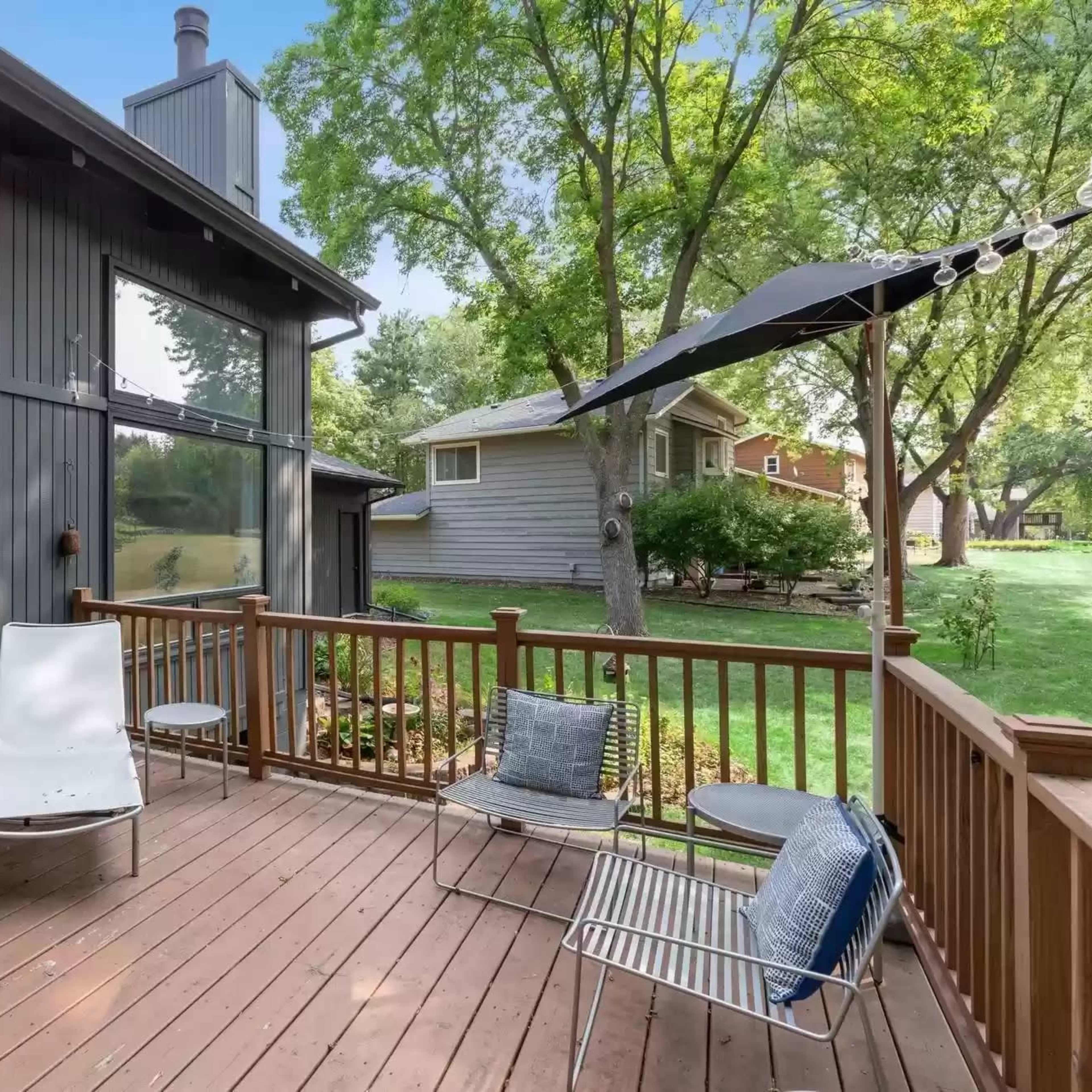 A wooden deck with metal chairs and a lounge chair, surrounded by trees and adjacent to a lawn with houses in the background.