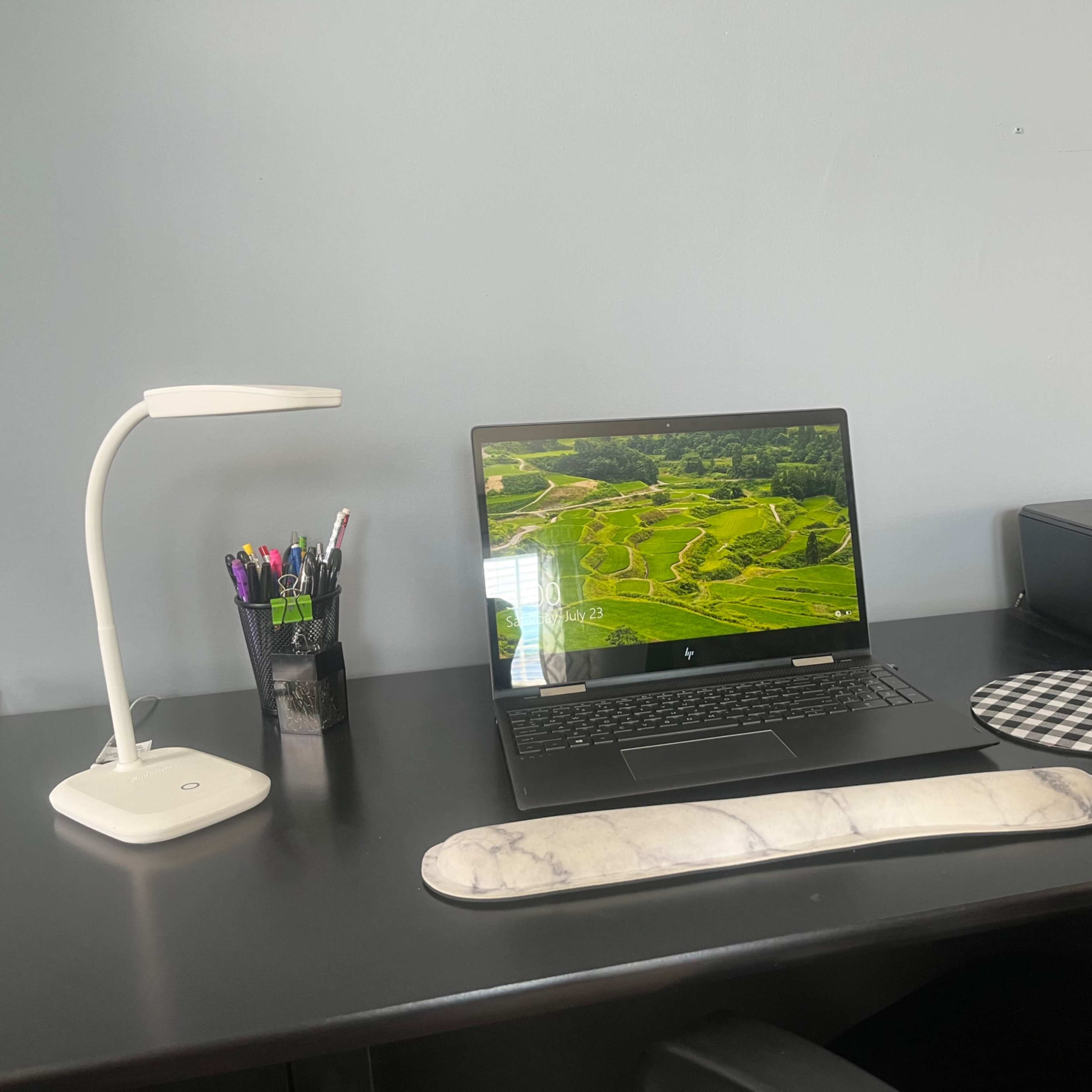 A laptop displaying a landscape image is placed on a black desk alongside a lamp, a printer, and various writing utensils in a cup.