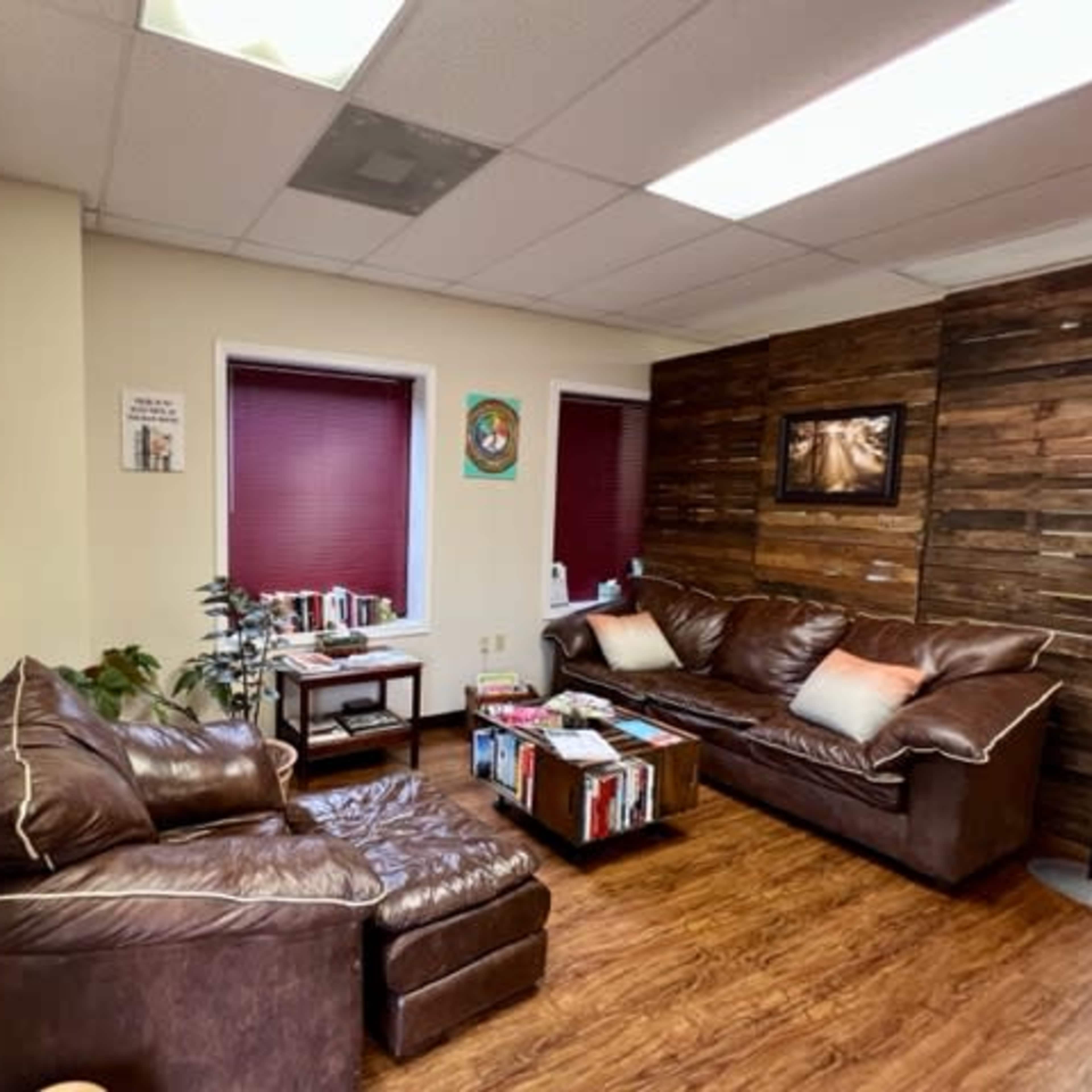 A cozy office waiting area with two leather sofas, a wooden accent wall, potted plants, and shelves filled with books.
