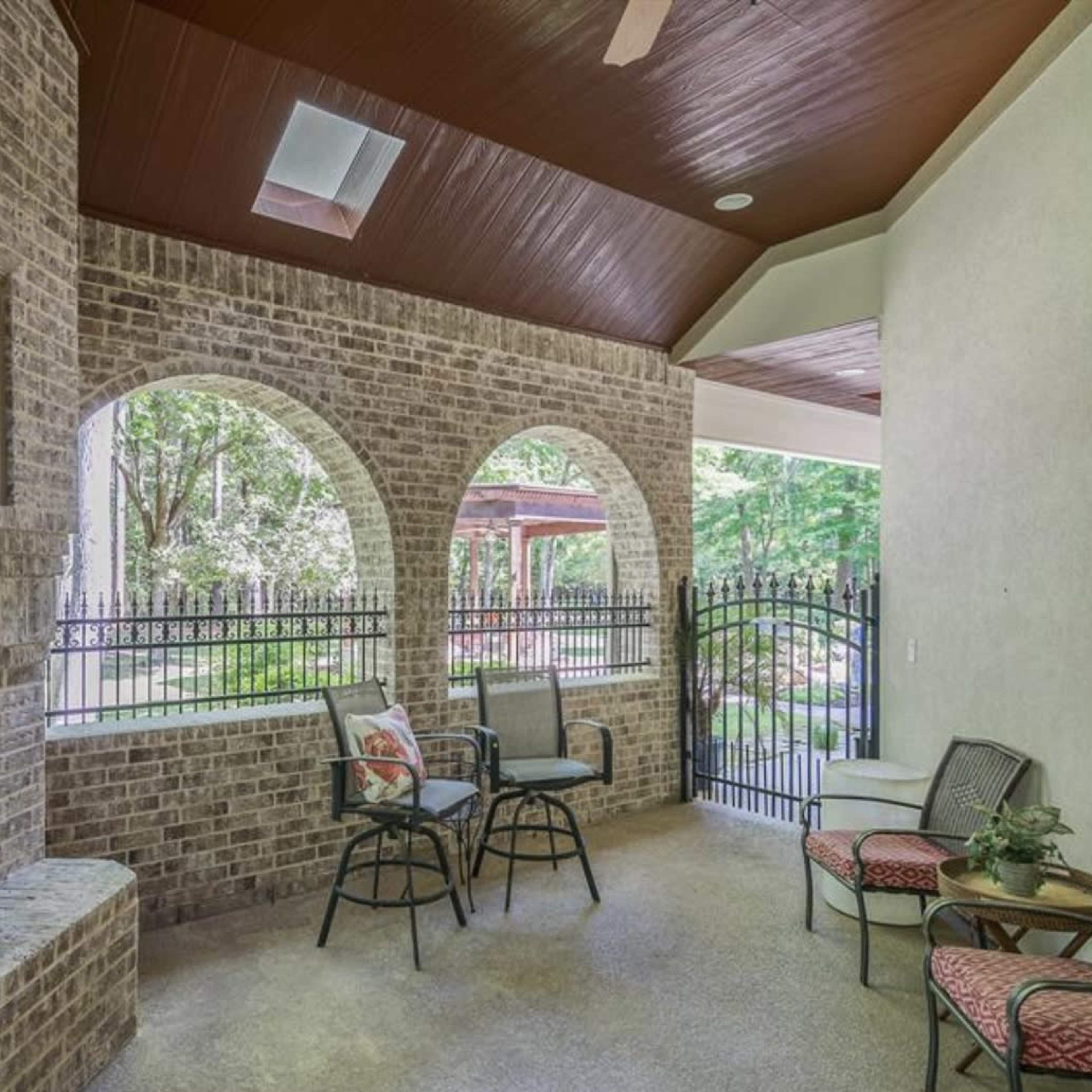 A covered porch area with brick walls, a fireplace, and several chairs arranged around a view of a yard with a fenced garden.
