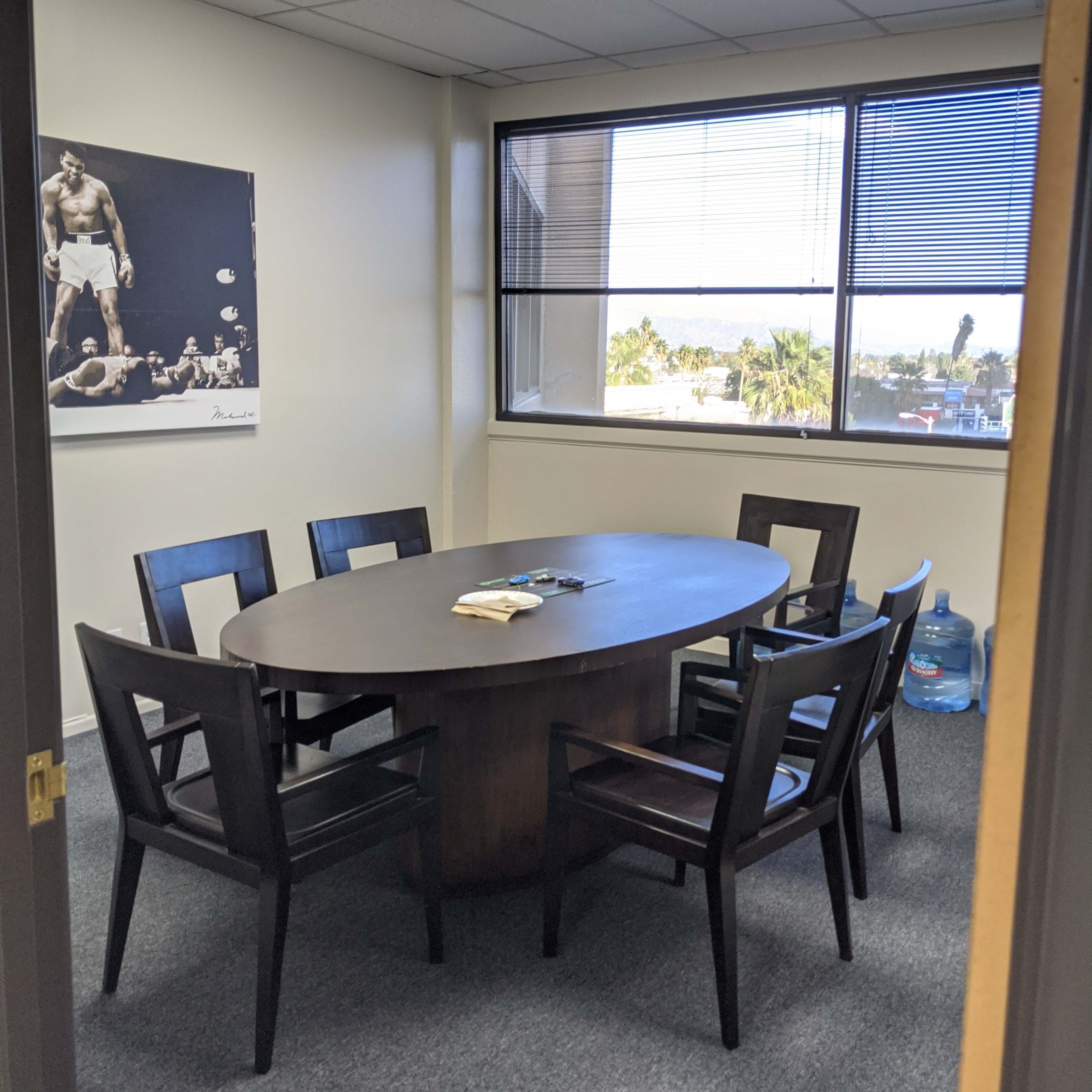 A conference room features an oval table surrounded by seven dark chairs, with a window displaying a view outside and a water dispenser in the corner.