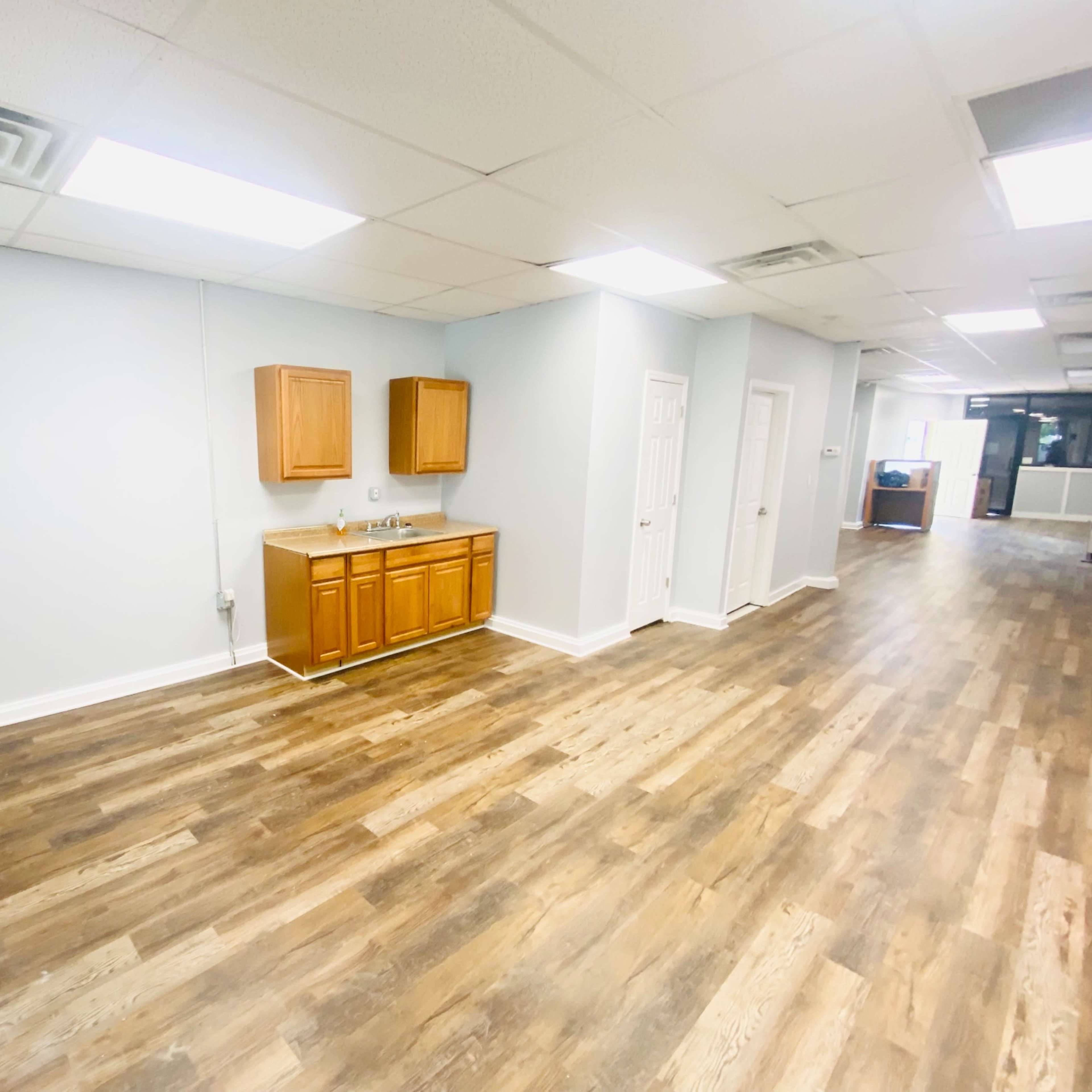A spacious room with wooden flooring and a kitchen area featuring brown cabinets against a light gray wall.
