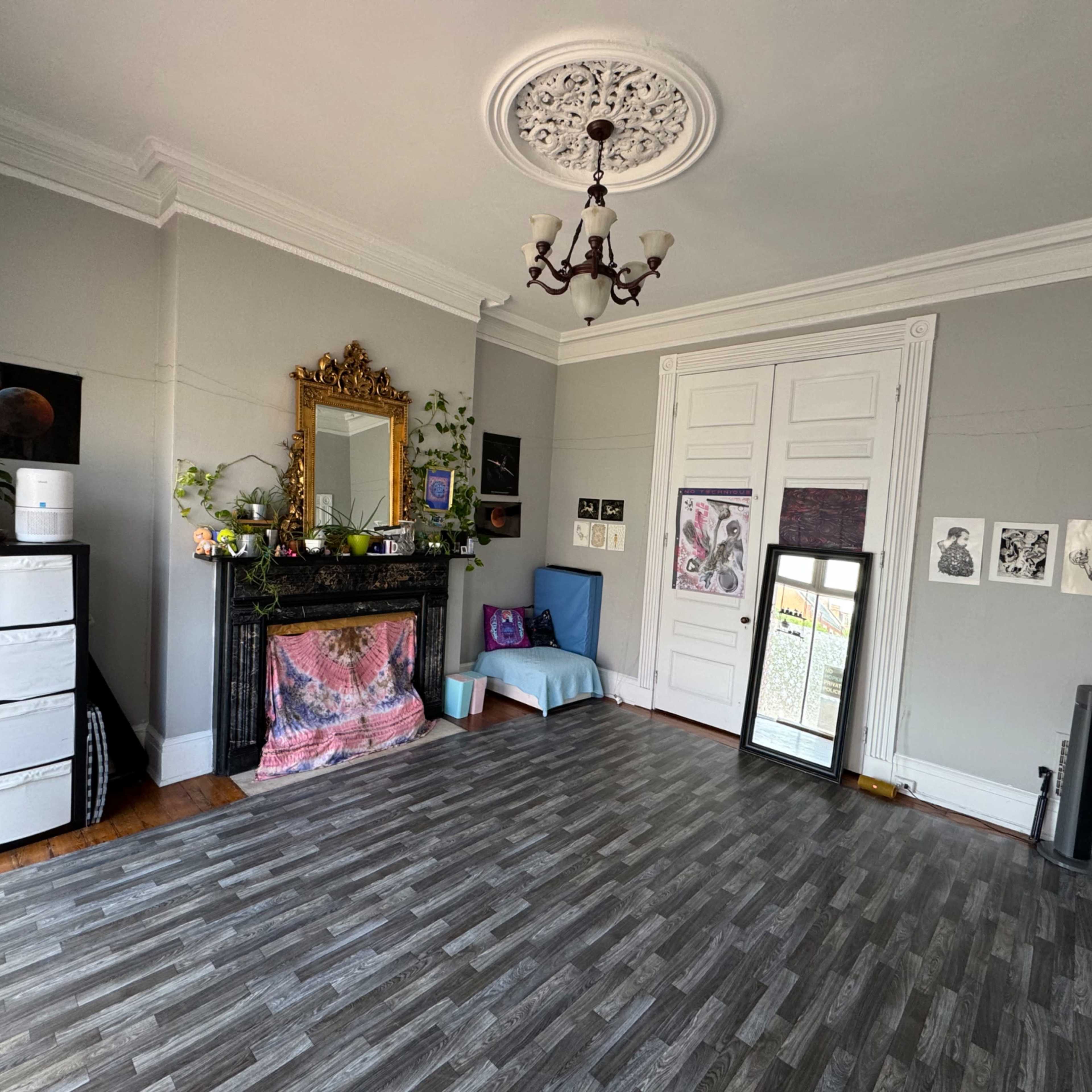 A spacious room features a black fireplace, a large mirror, various plants, and artwork hanging on the walls, with a blue couch against one side and a gray wood-like floor.