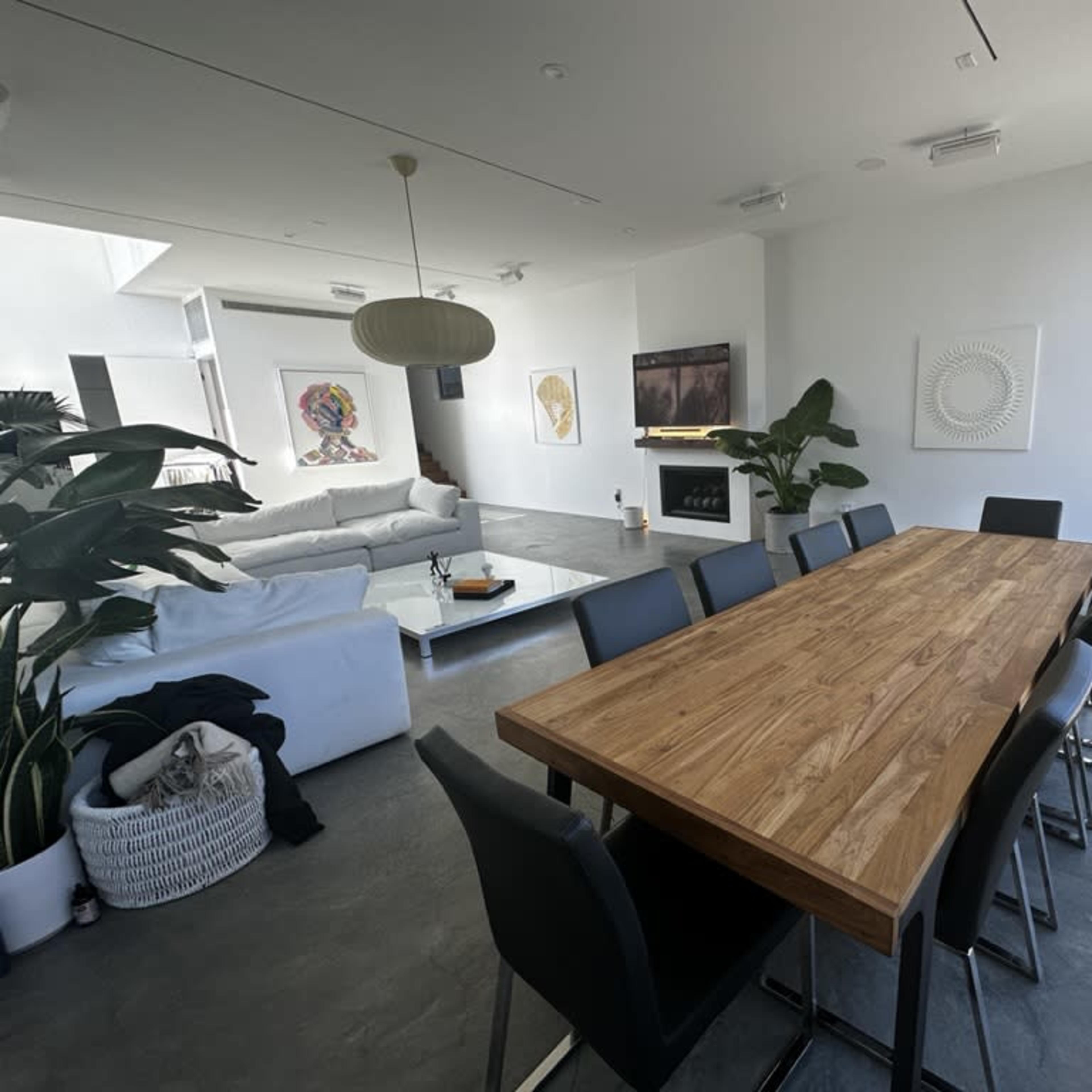A spacious dining area features a long wooden table surrounded by black chairs, overlooking a modern living space with a white couch and large windows.