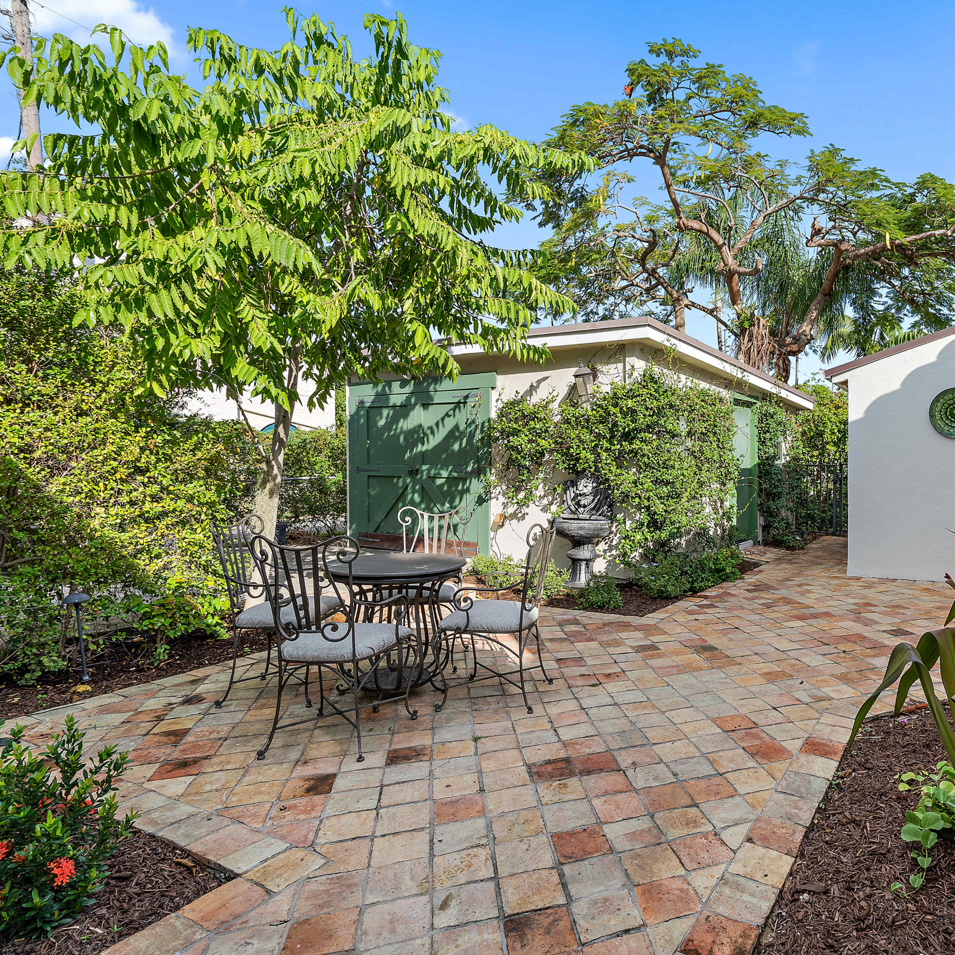 A paved garden area features a table and chairs surrounded by lush greenery and flowering plants.