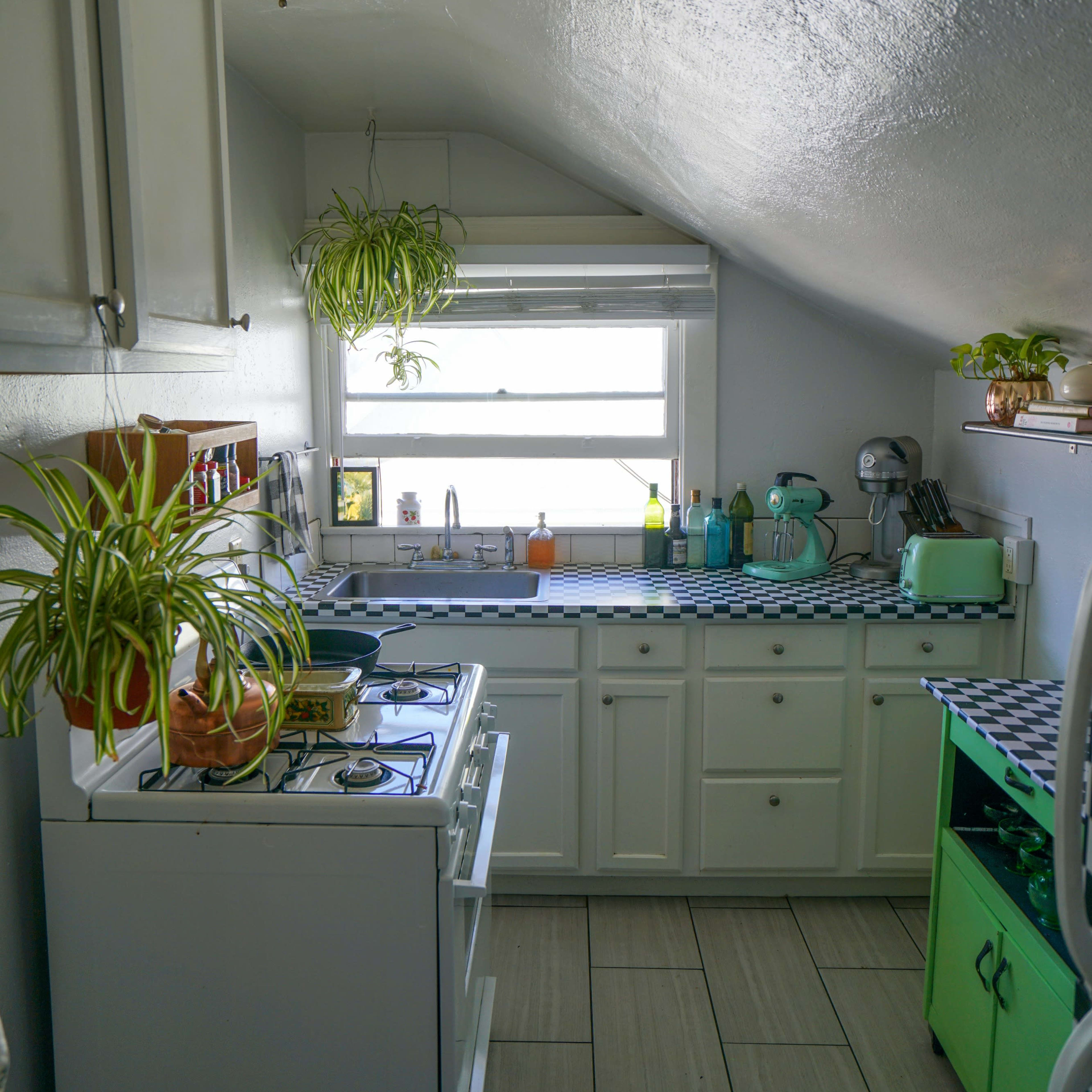 A compact kitchen with slanted ceilings, featuring a white stove, white cabinets, and a green fridge, along with houseplants on the countertop and a window providing natural light.