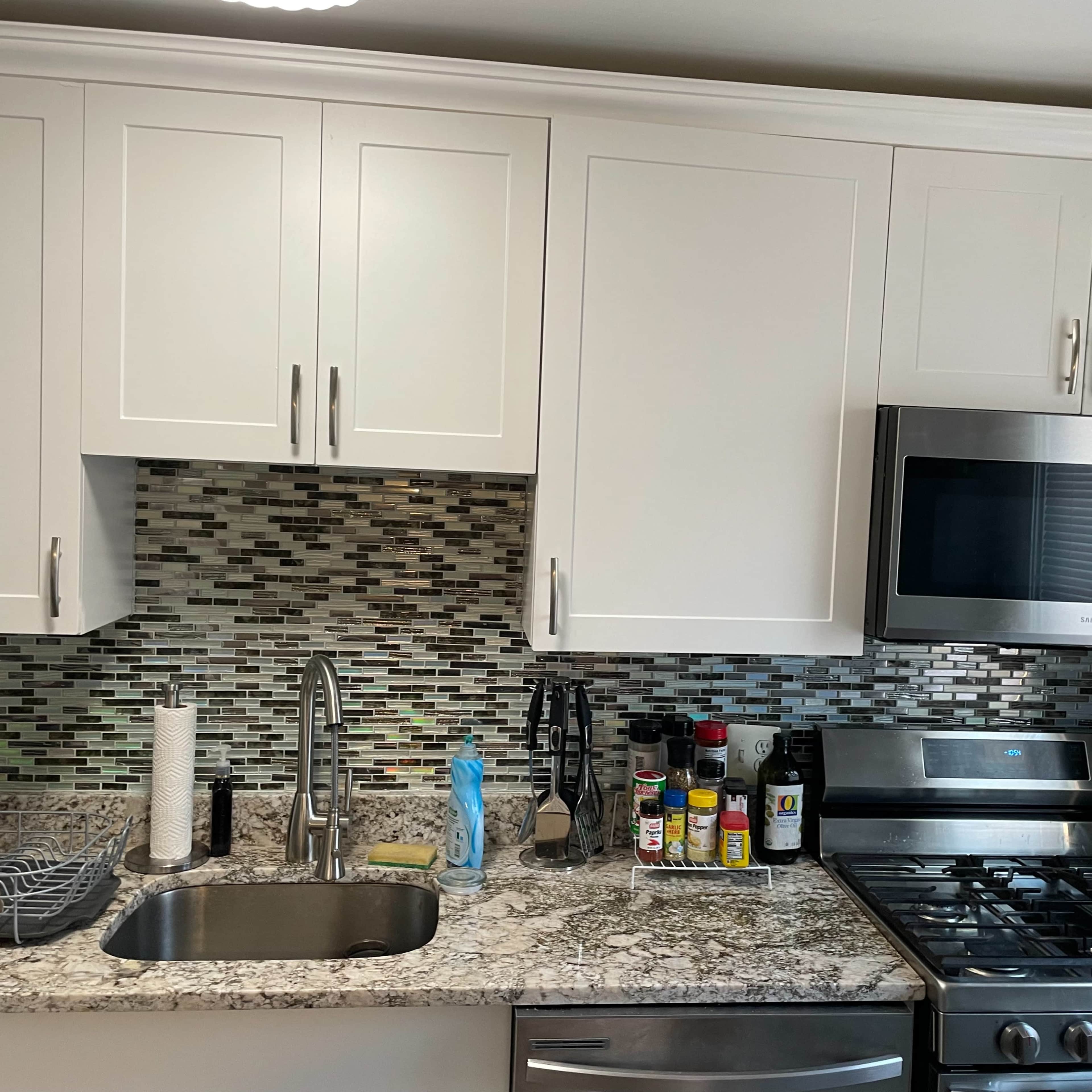 A modern kitchen with white cabinets, a stainless steel microwave, a gas stove, and a granite countertop featuring a sink and various condiments.