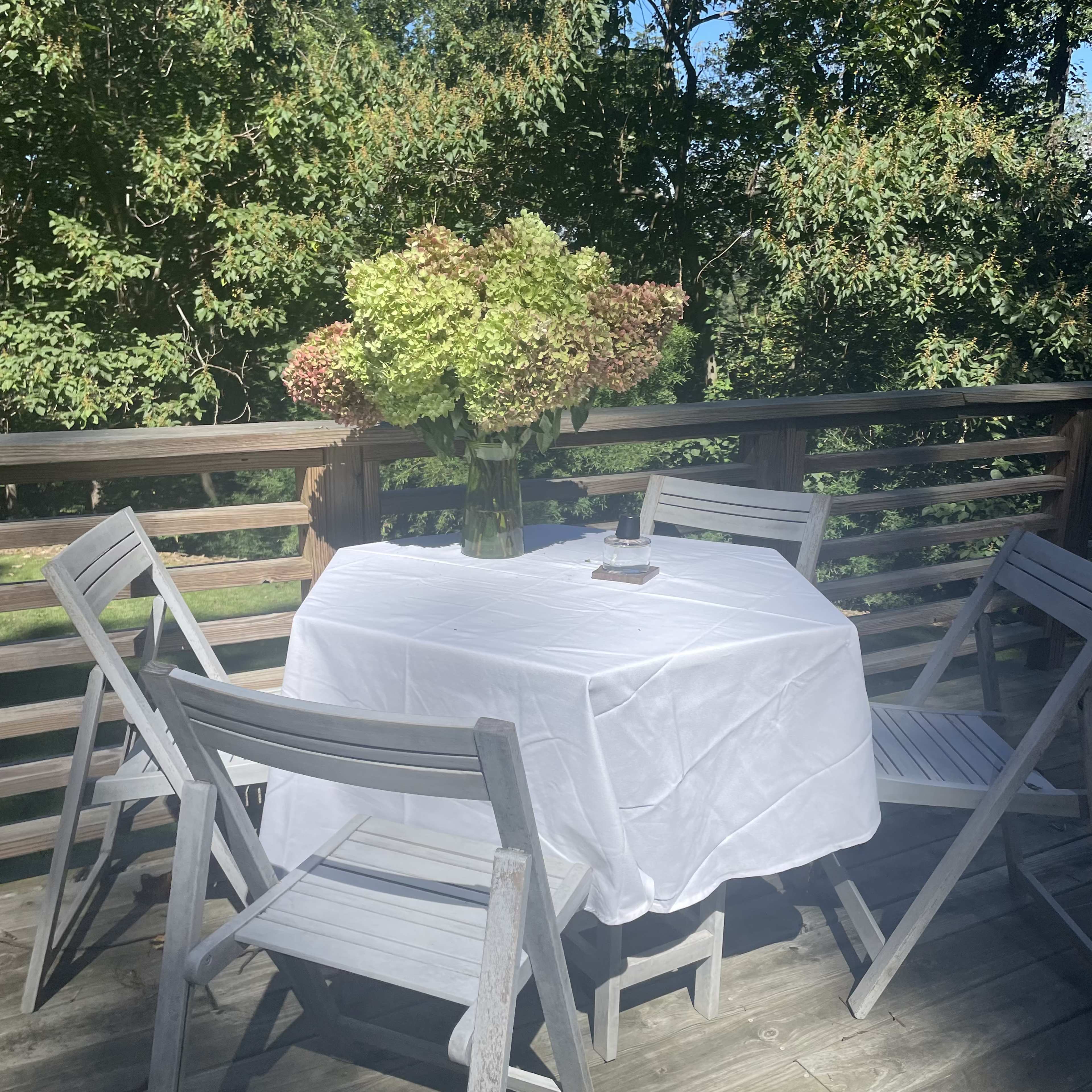 A round table covered with a white tablecloth is set on a wooden deck surrounded by greenery, with a large bouquet of hydrangeas in a vase.
