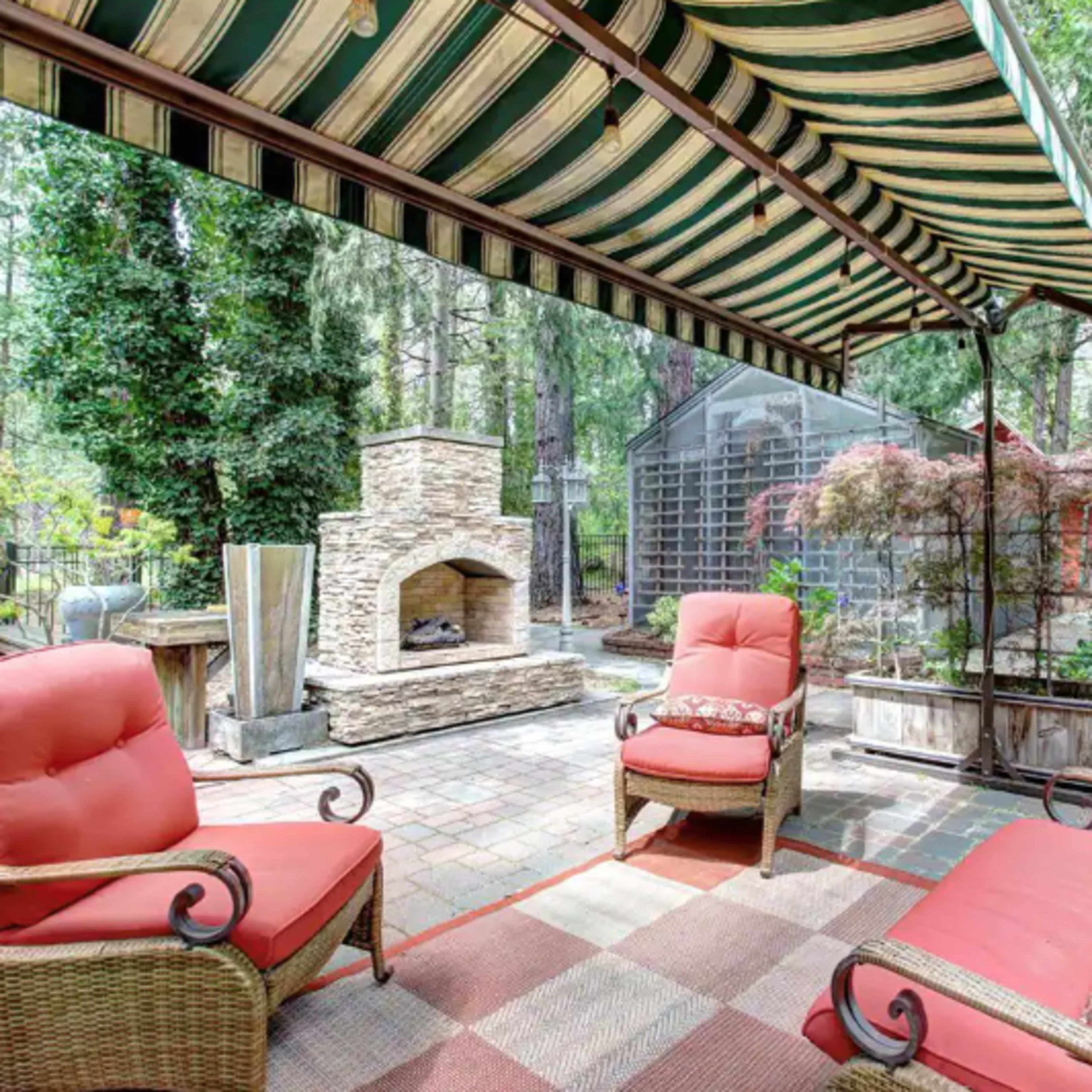 A patio area features two red chairs, a stone fireplace, and a striped awning, surrounded by lush greenery.