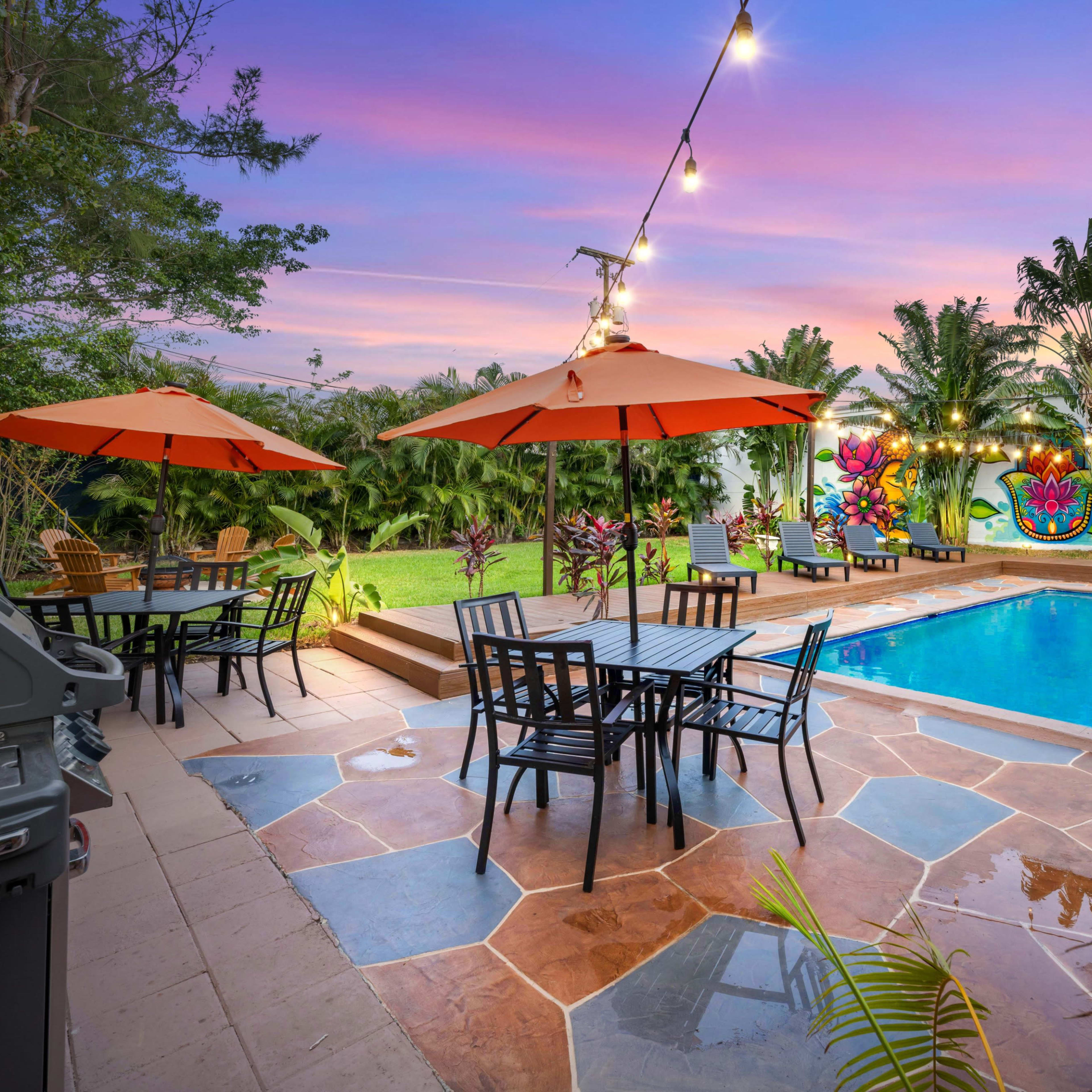 A backyard with a swimming pool, surrounded by patio furniture under orange umbrellas, and a vibrant mural on the wall.