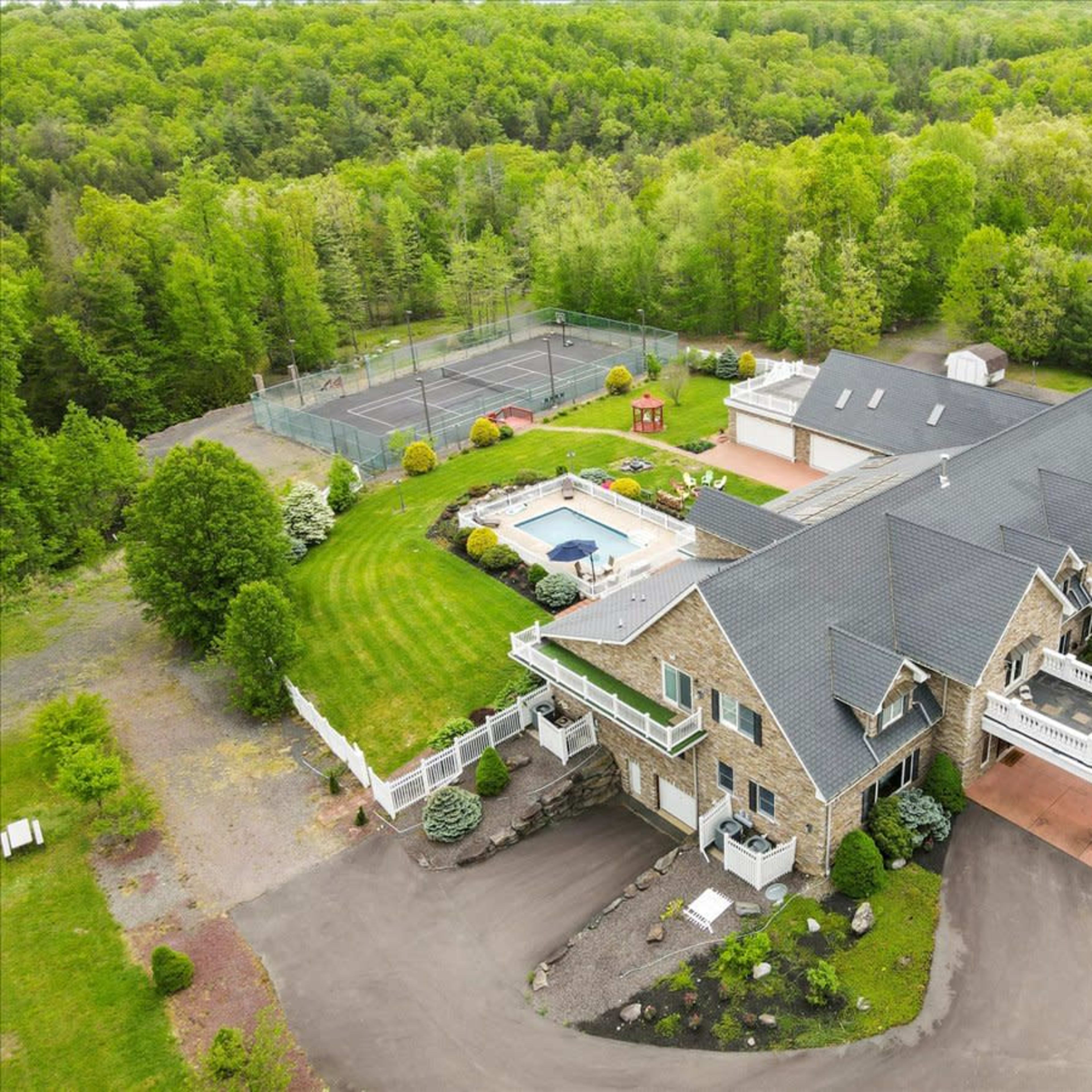 A large stone house with a swimming pool and tennis court surrounded by lush greenery.