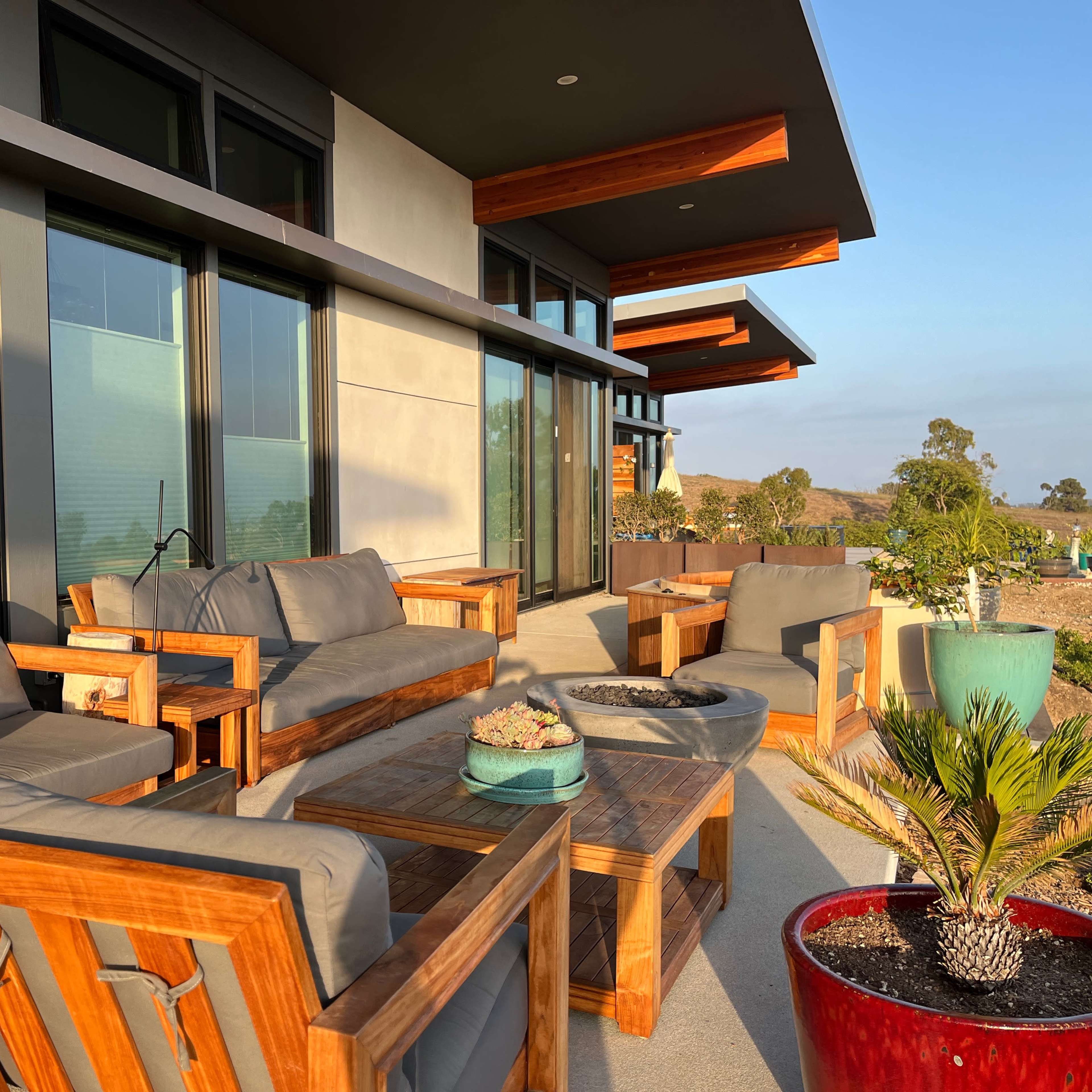 A modern outdoor seating area with wooden furniture, a fire pit, and a view of landscaped greenery in the background.