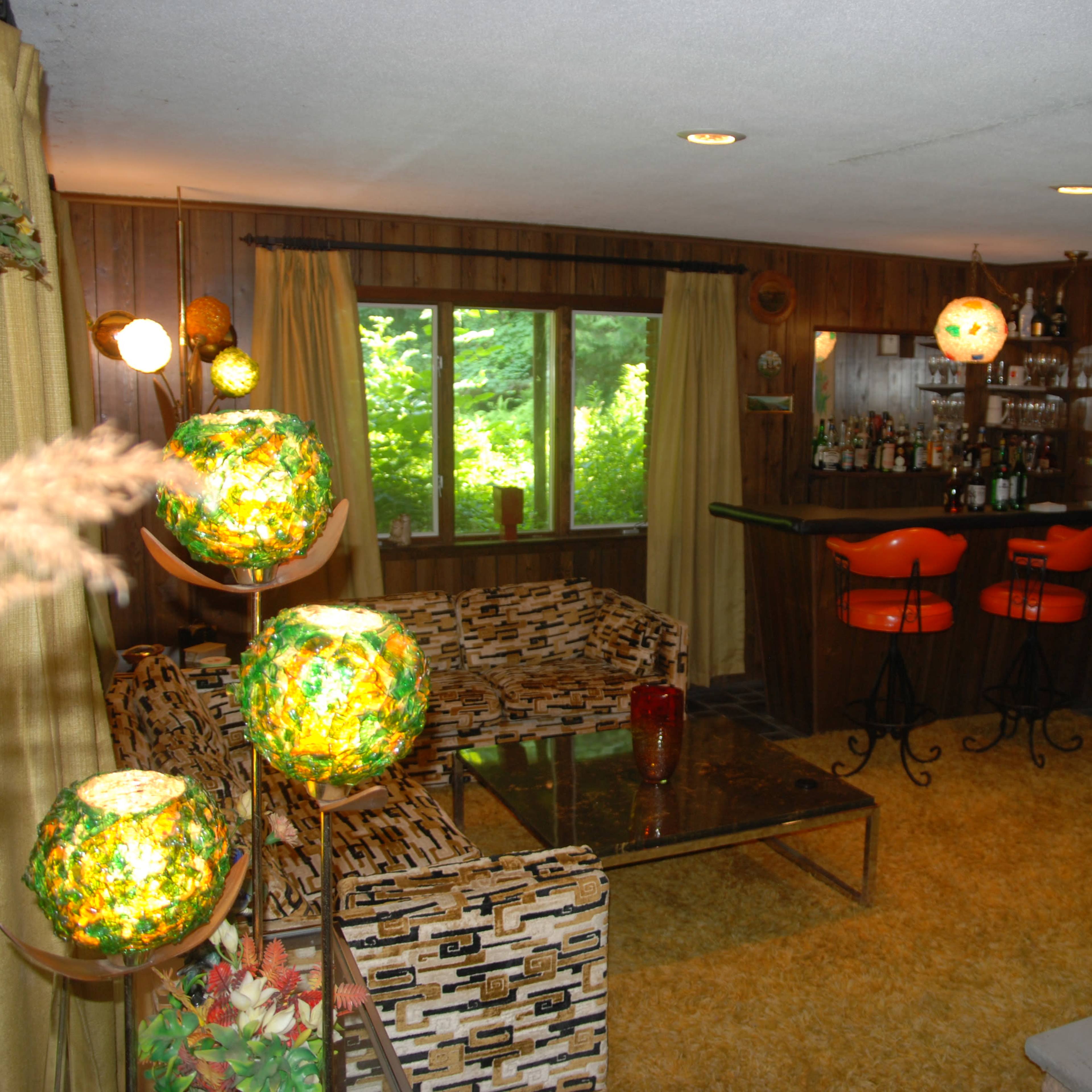 A retro-styled living room with wooden walls, a bar area, and orange stools, featuring a patterned couch and decorative lighting.