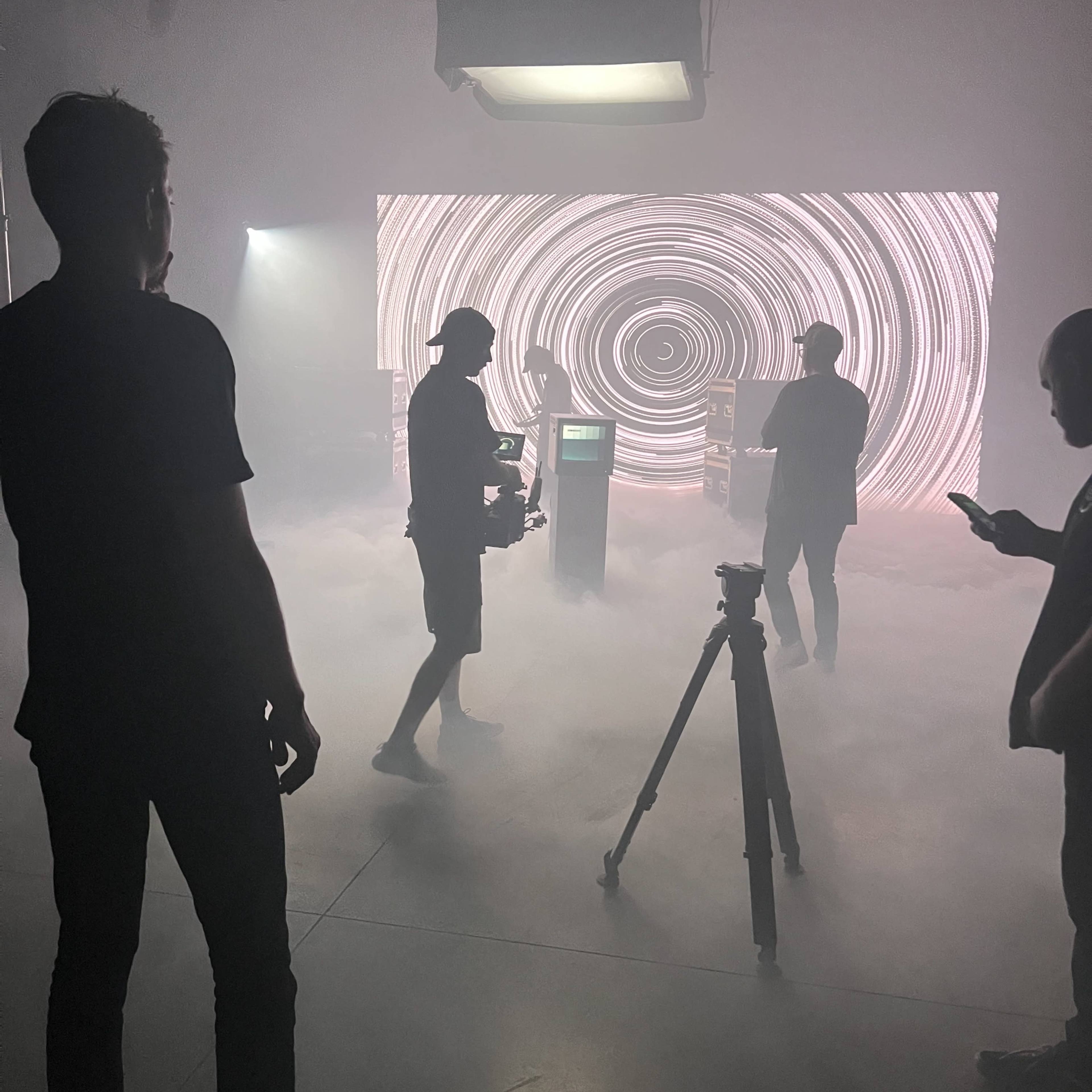 A group of individuals sets up a scene in a fog-filled space with a swirling light display in the background.