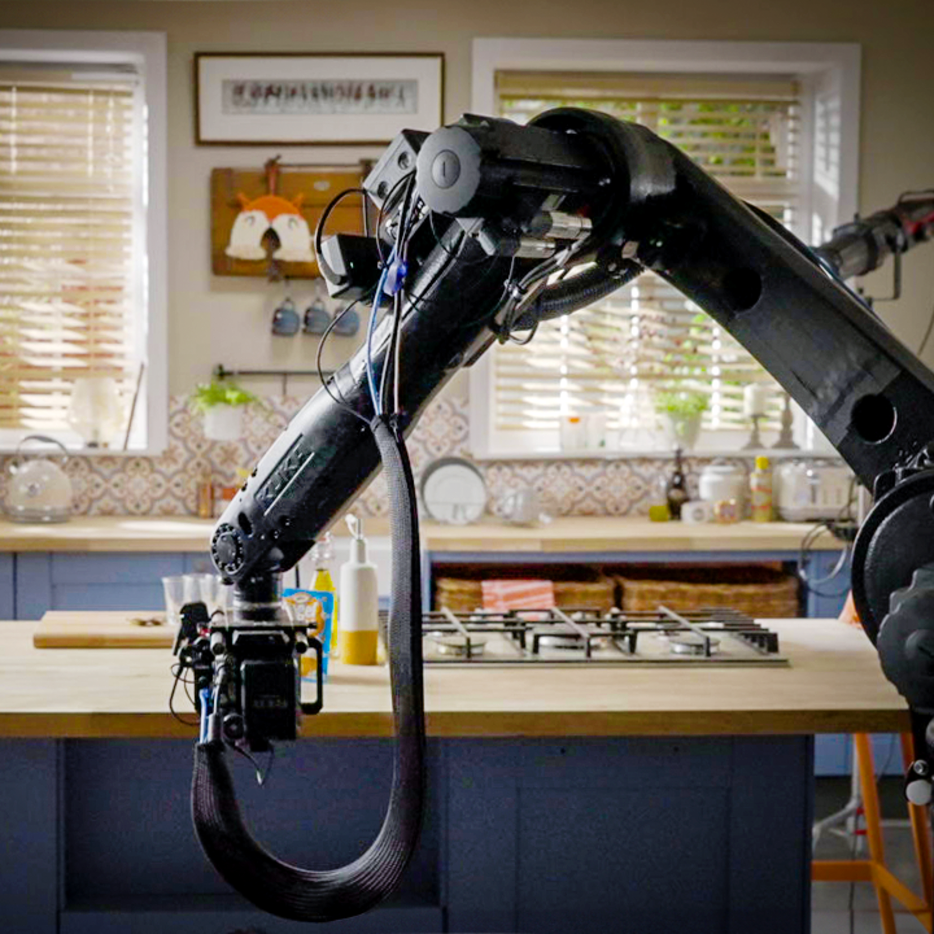 A robotic arm is positioned above a kitchen counter with a stovetop, surrounded by natural light and various kitchen items.