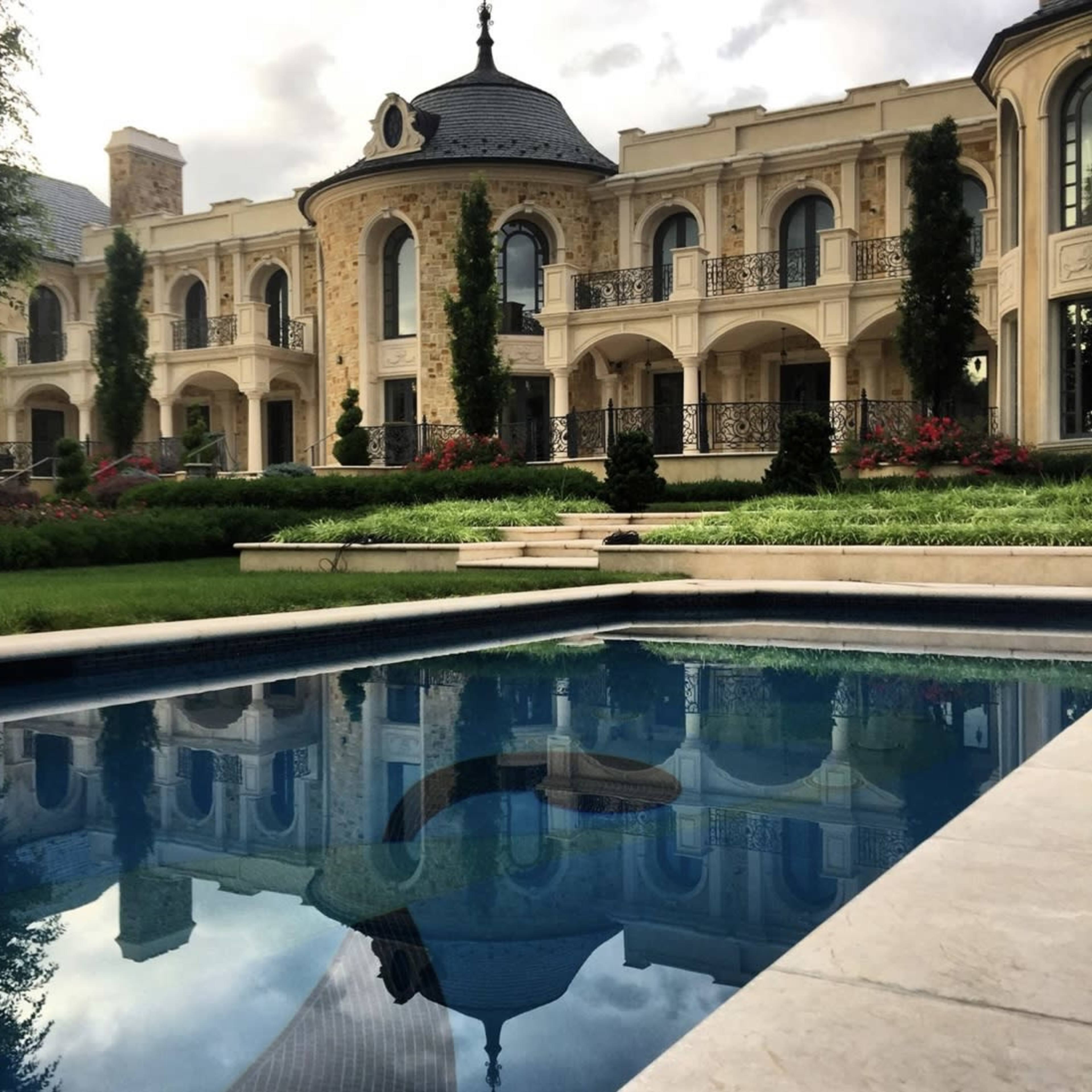 A large, elegant mansion with ornate architecture is reflected in a serene swimming pool, surrounded by lush landscaping and manicured gardens.