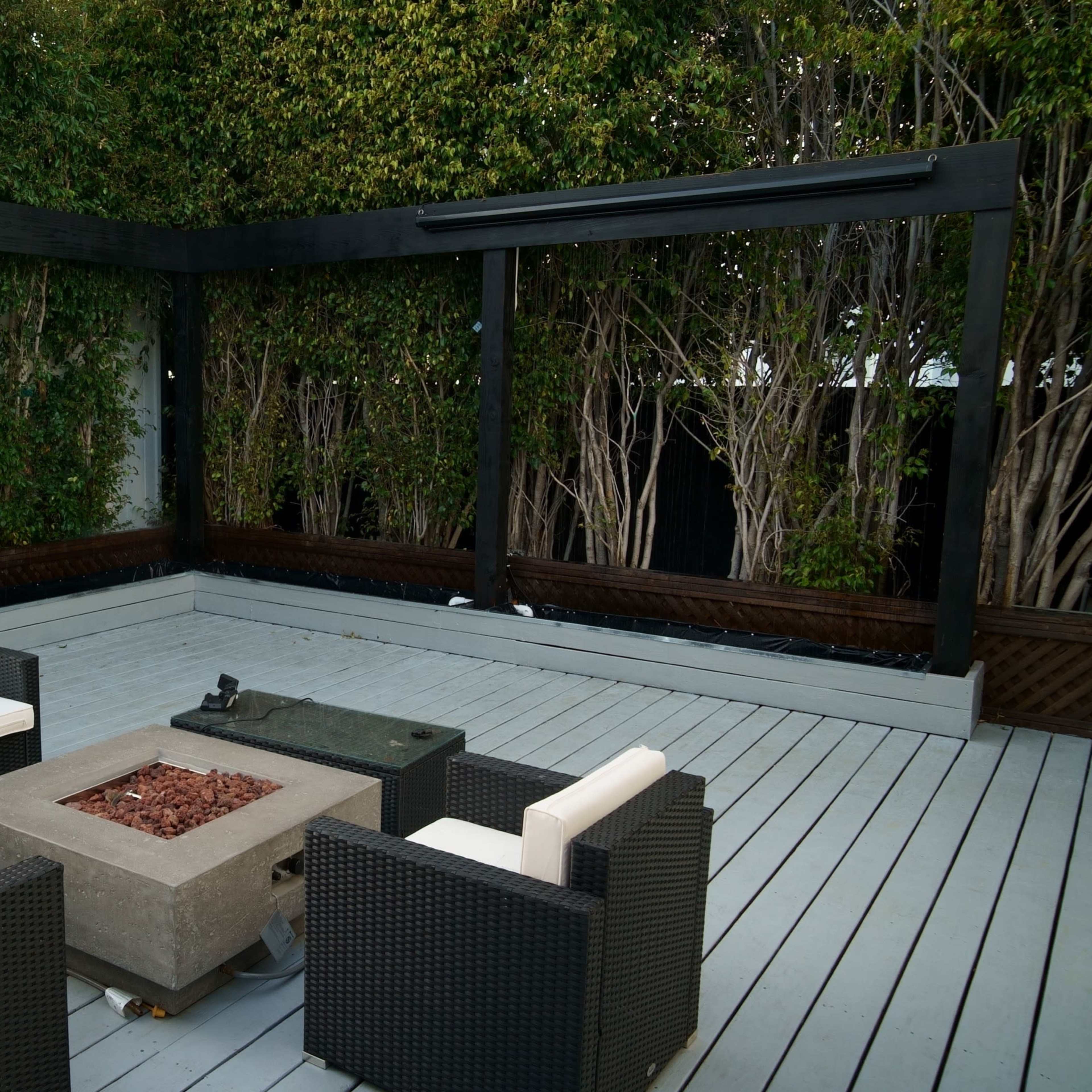 A patio area with a fire pit surrounded by four chairs, enclosed by tall greenery and a wooden frame.