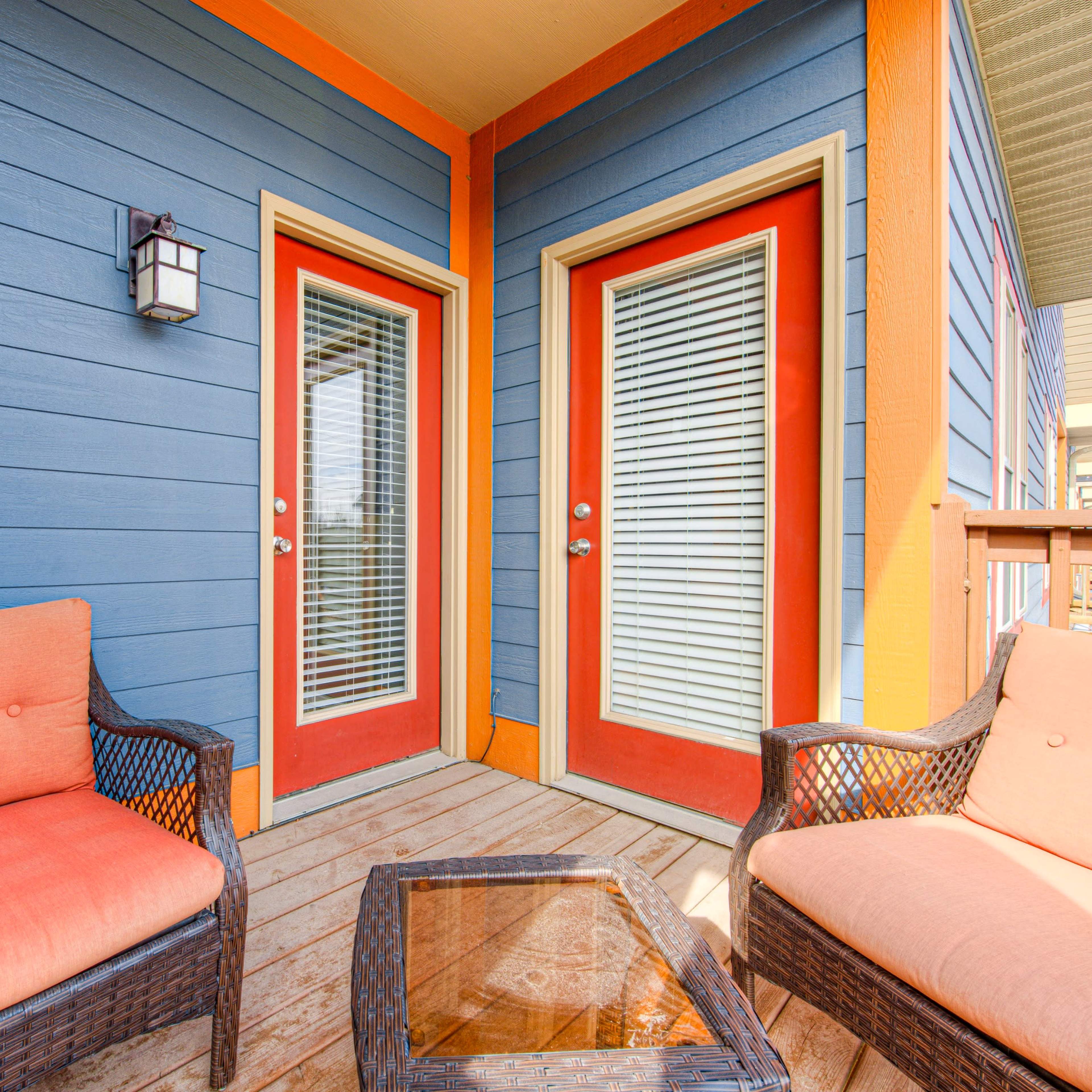 Two orange-cushioned chairs and a glass-top table are positioned on a wooden deck in front of two glass-paneled doors against a blue and orange exterior.