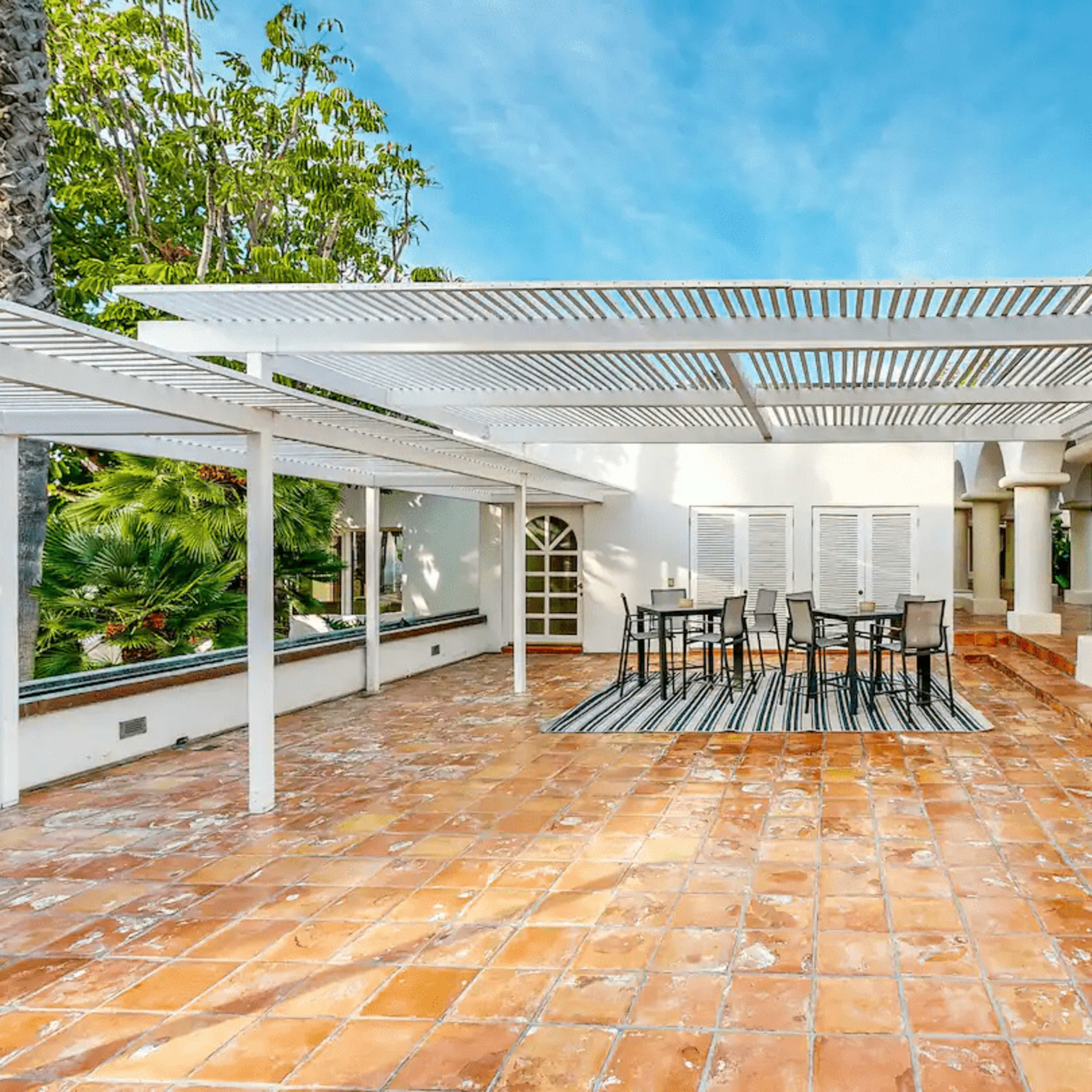 A spacious outdoor patio with a tiled floor, covered seating area, and white pergola, surrounded by tropical greenery.