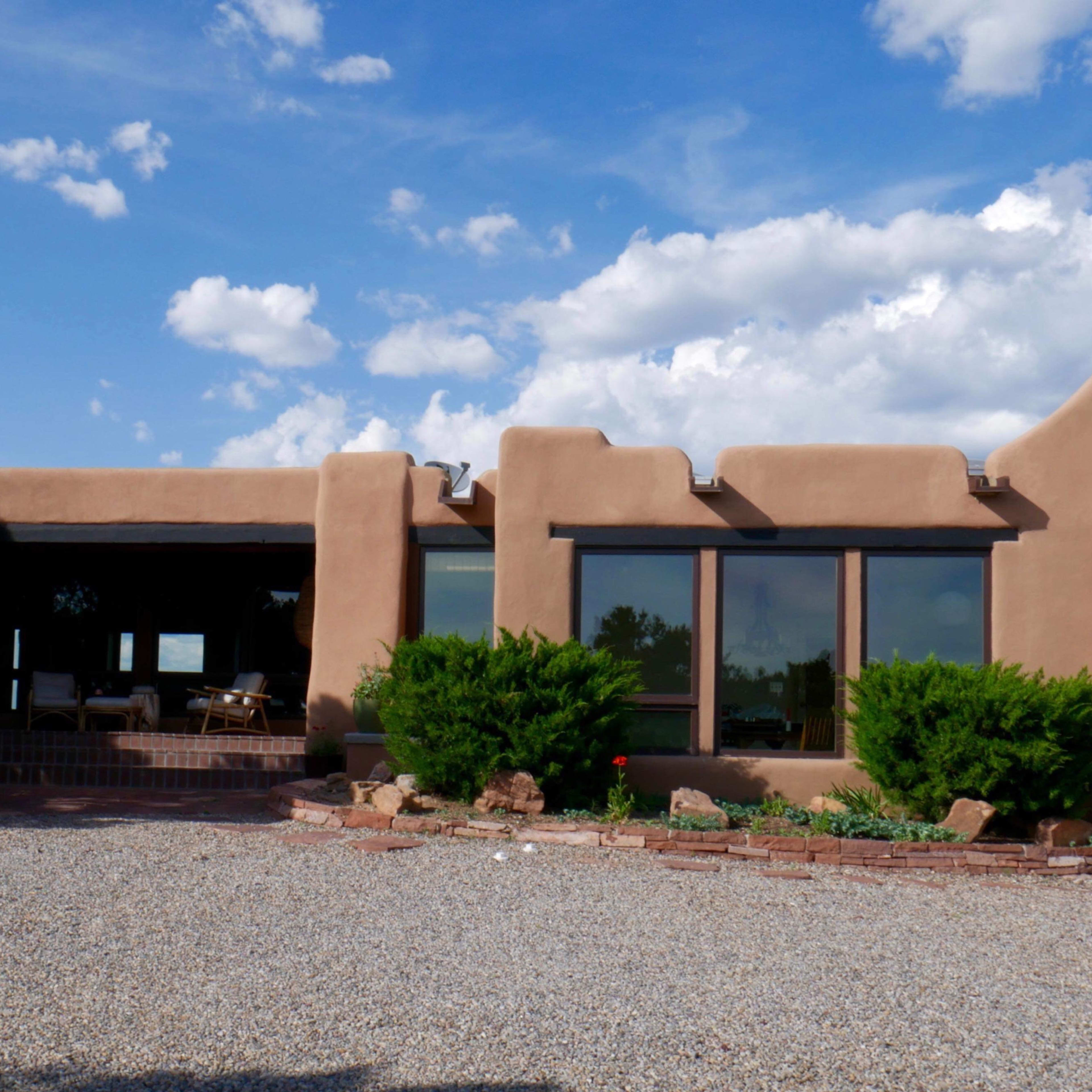 A single-story adobe-style house with large windows, set against a backdrop of blue sky and scattered clouds.