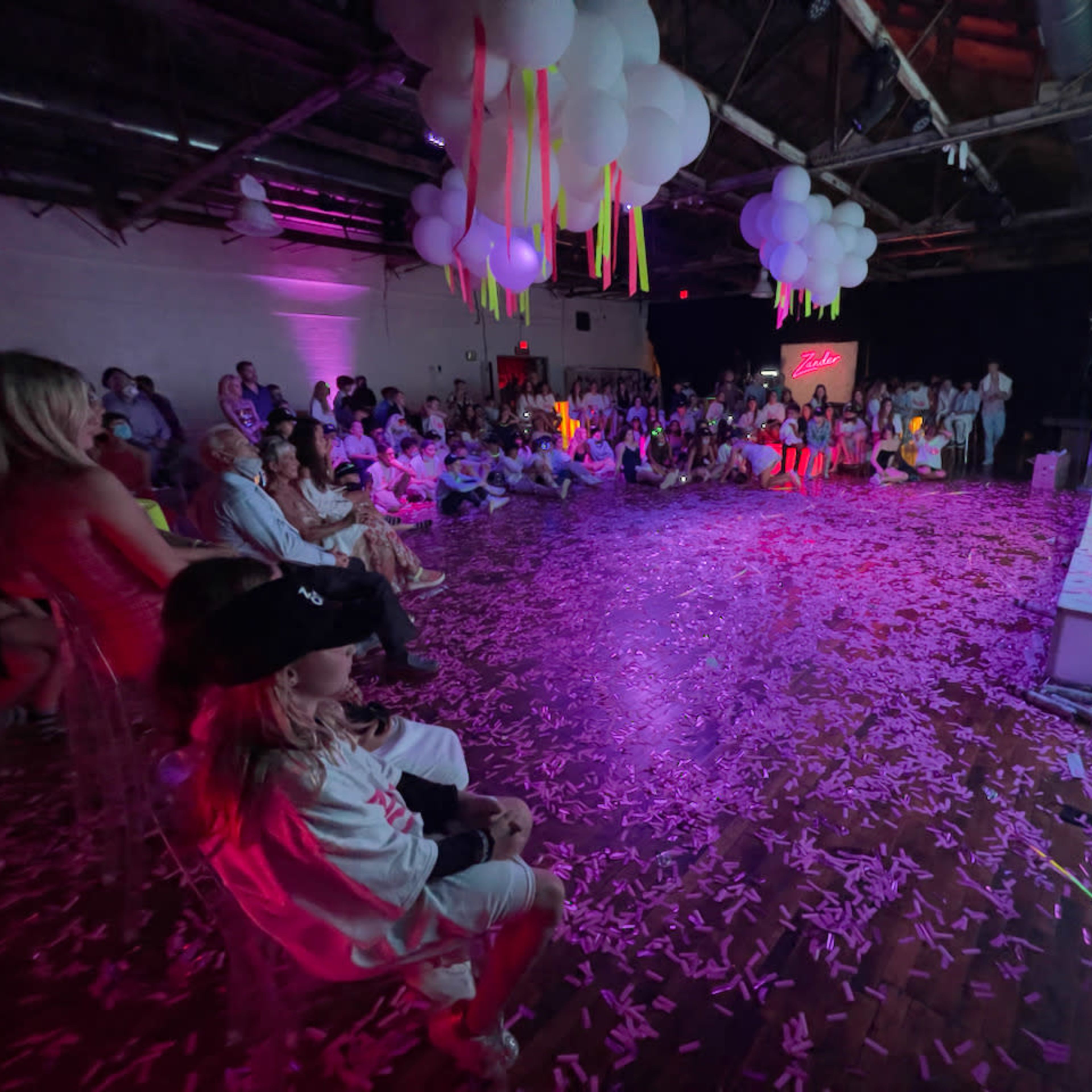 A crowd gathers in a spacious venue decorated with balloons and colorful streamers, watching a large screen while confetti covers the floor.