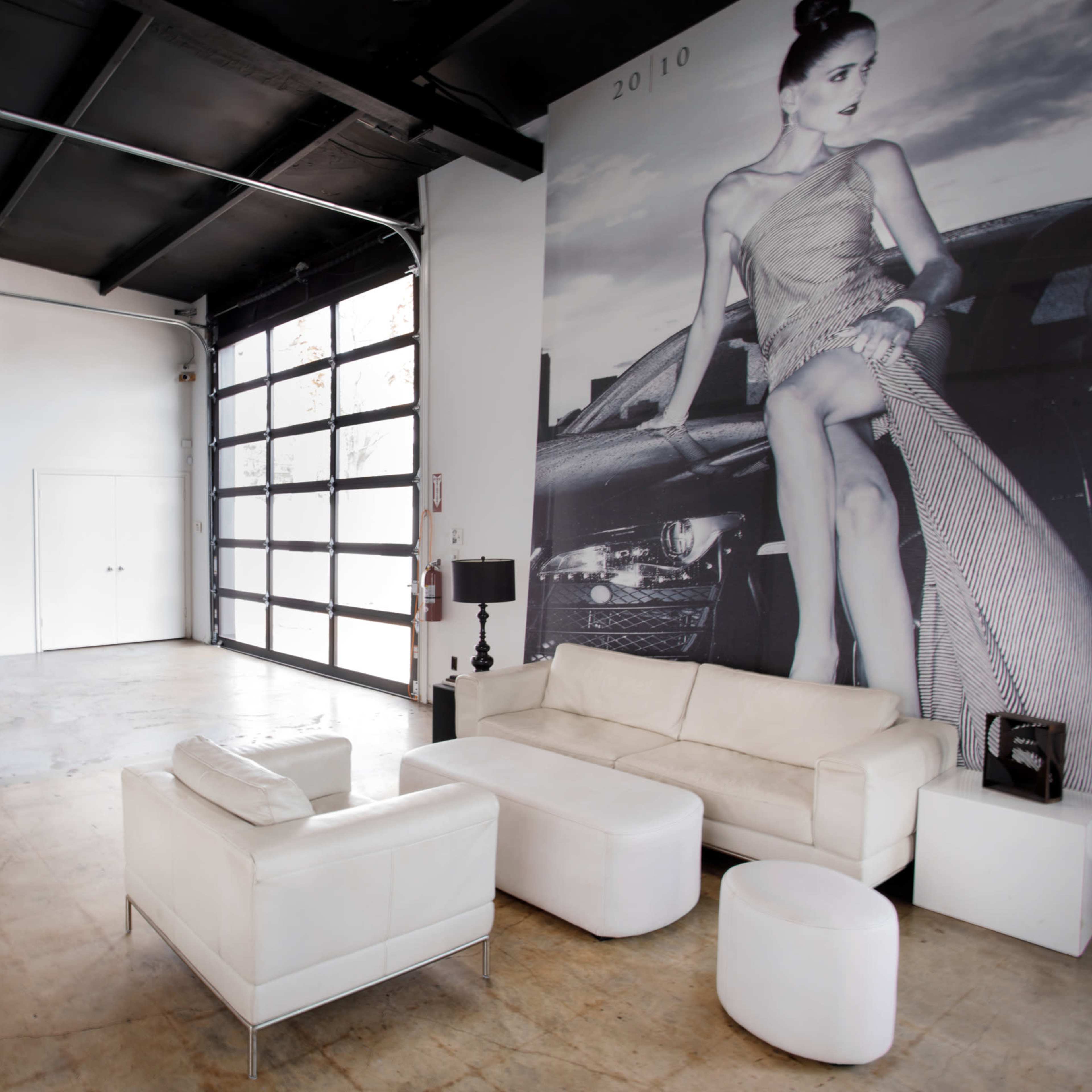 A minimalist interior with a white sofa set and large glass doors featuring a large wall mural of a woman beside a car.