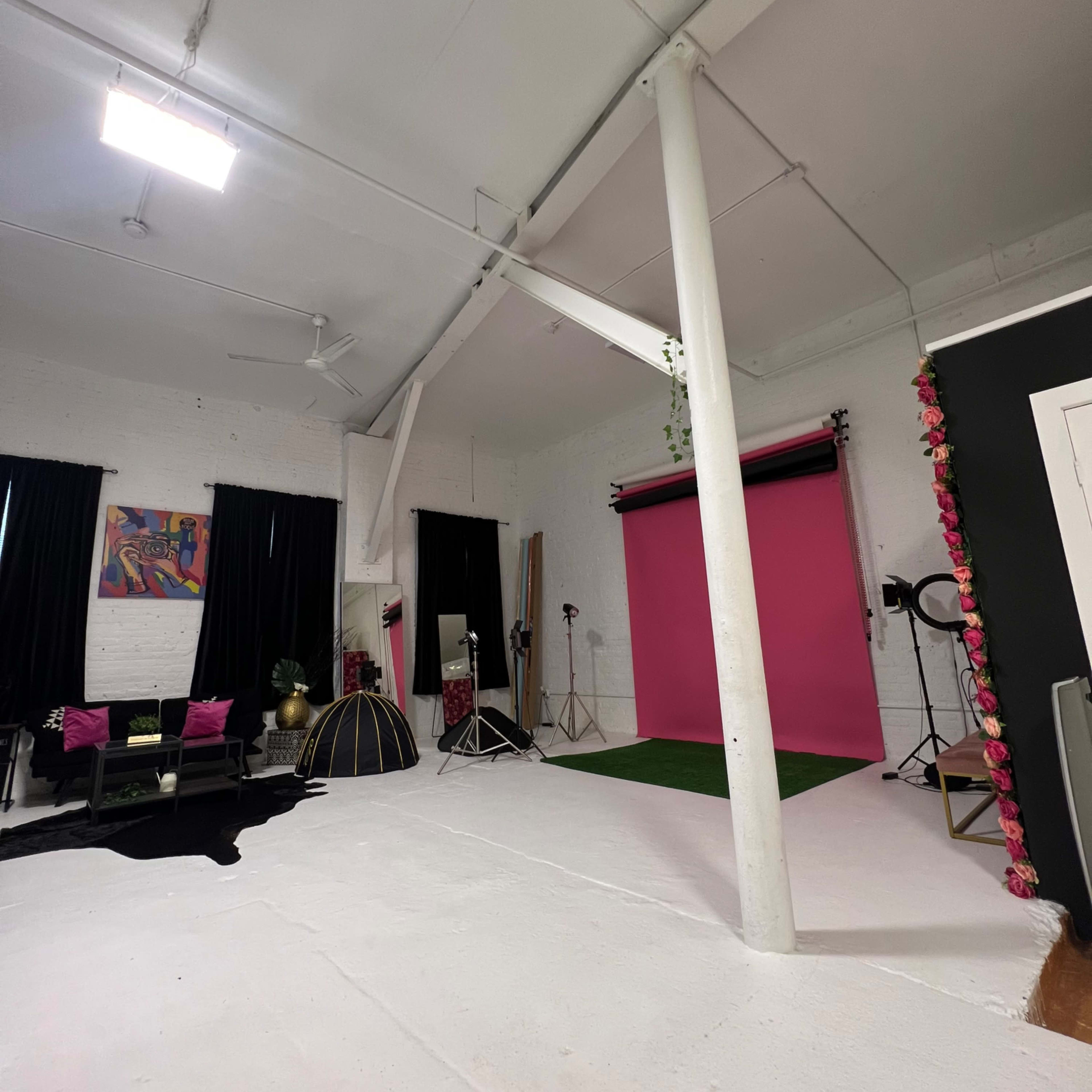 A photography studio with a pink backdrop, green carpet, and various lighting equipment set up, alongside black curtains and furniture.