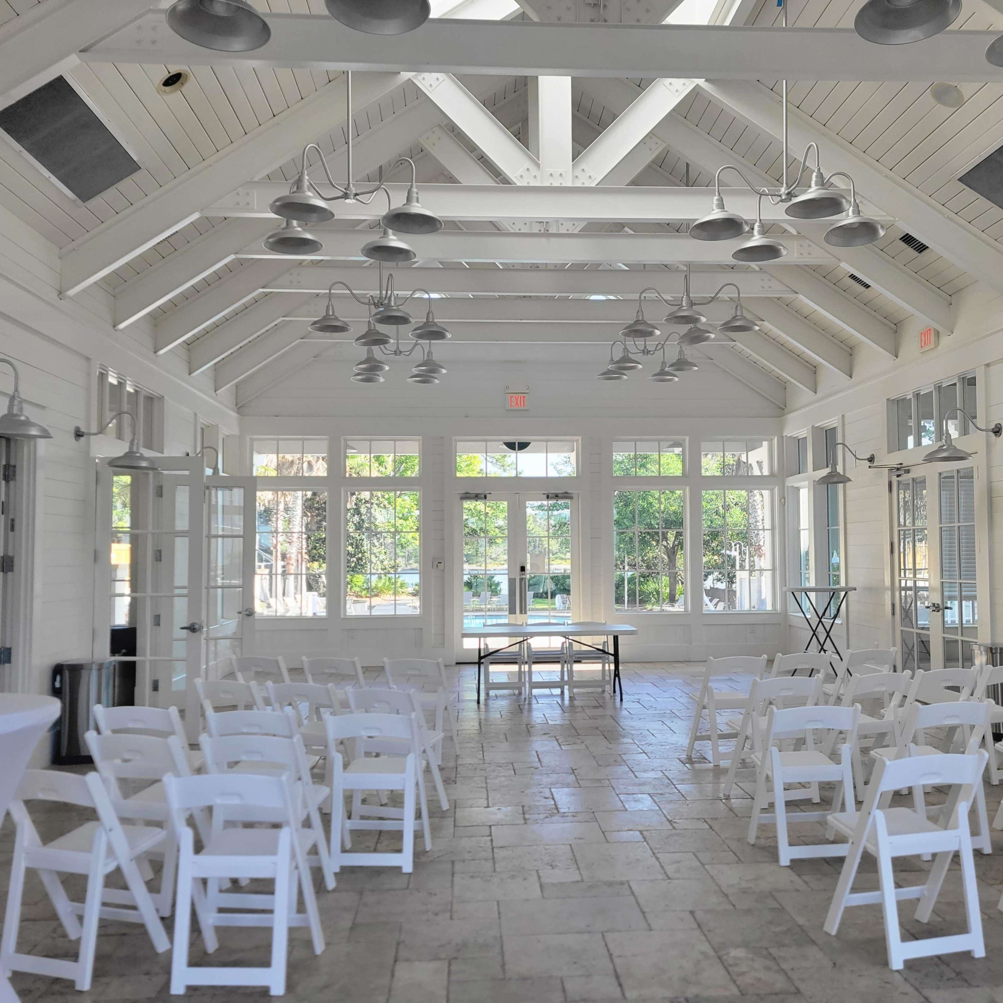 A well-lit, white indoor venue features rows of white chairs facing a table, with large windows providing a view of the outdoors.