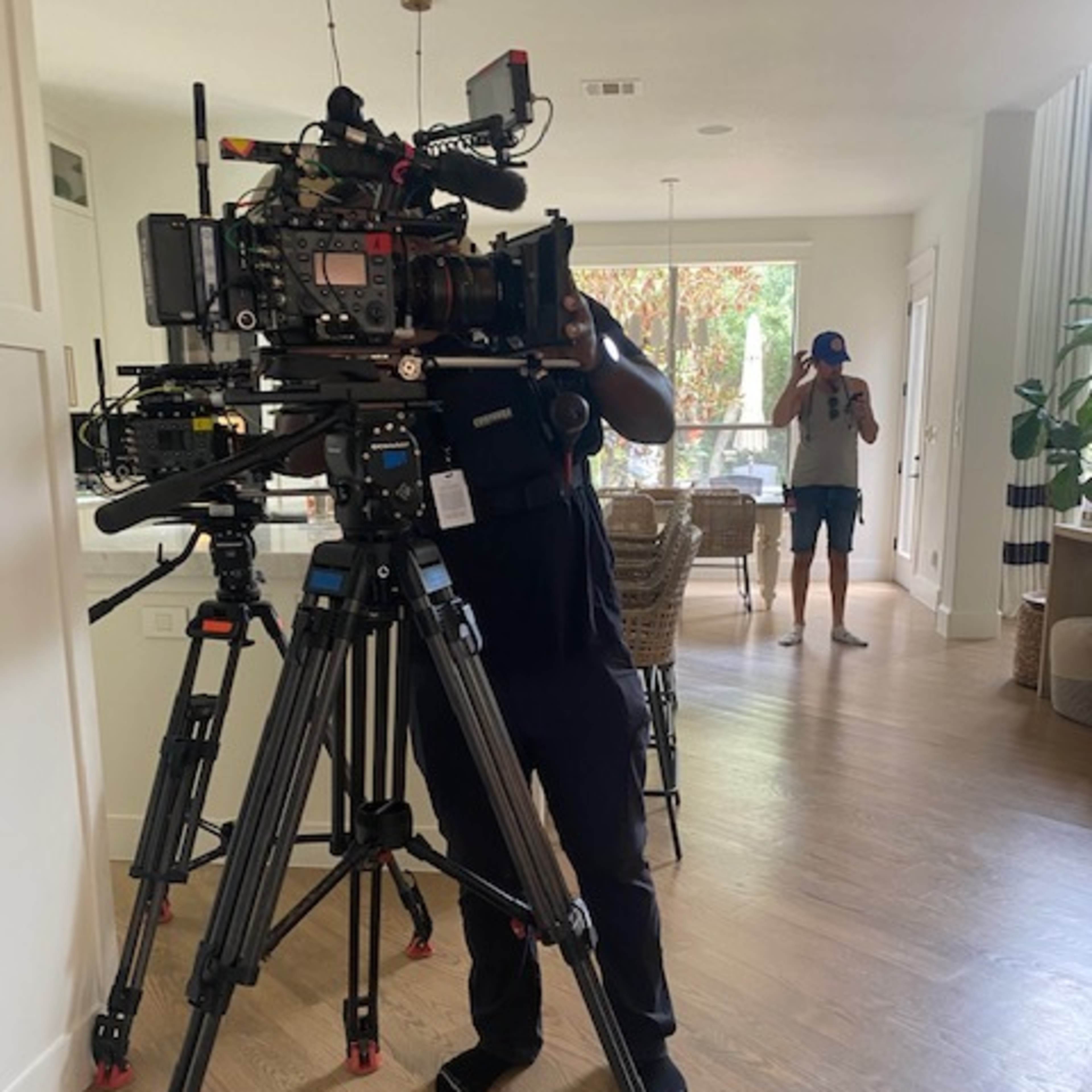 A camera operator sets up a film camera on a tripod in a well-lit room while a person in the background holds a phone.