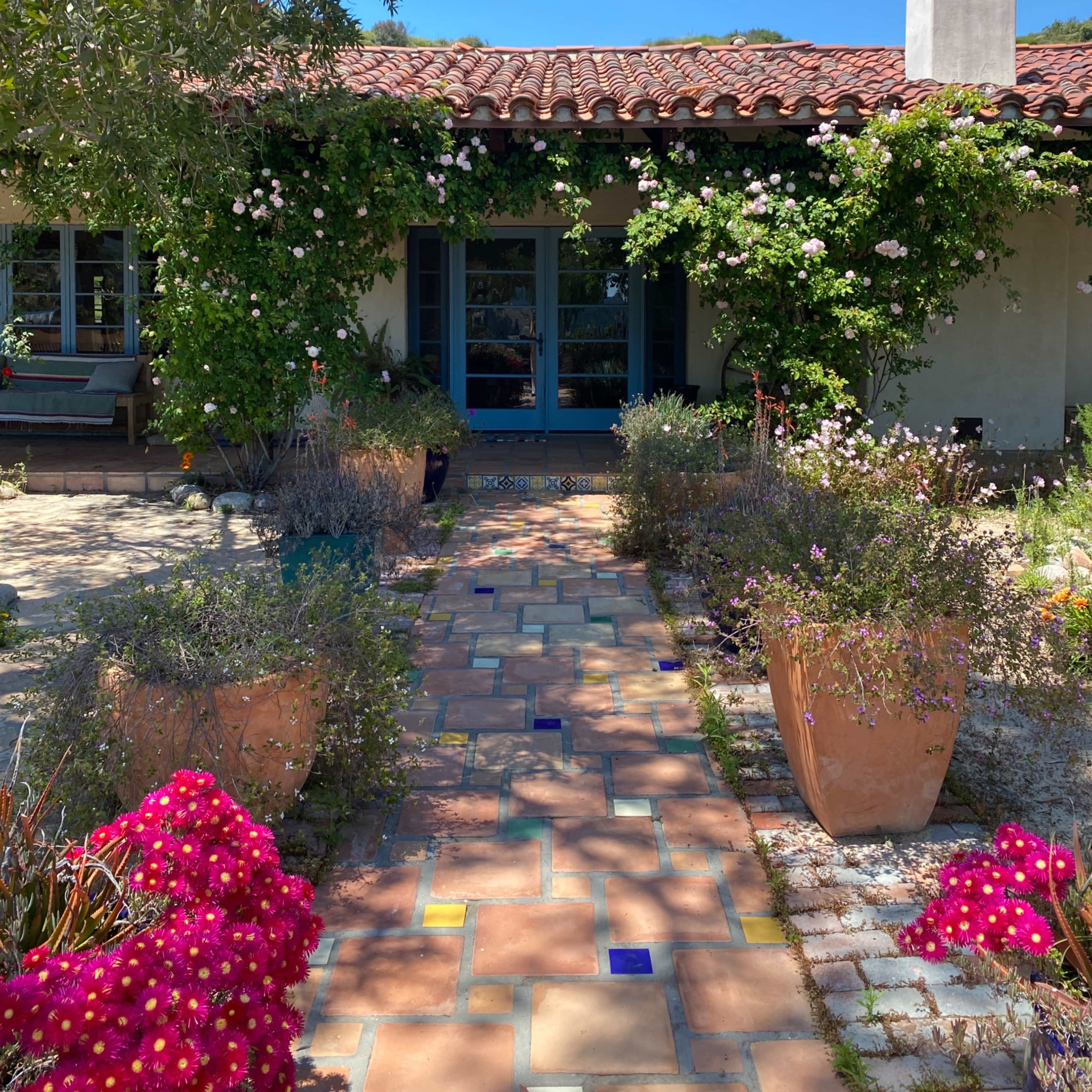 A path made of colorful tiles leads to a house adorned with climbing roses and surrounded by vibrant flowerpots.