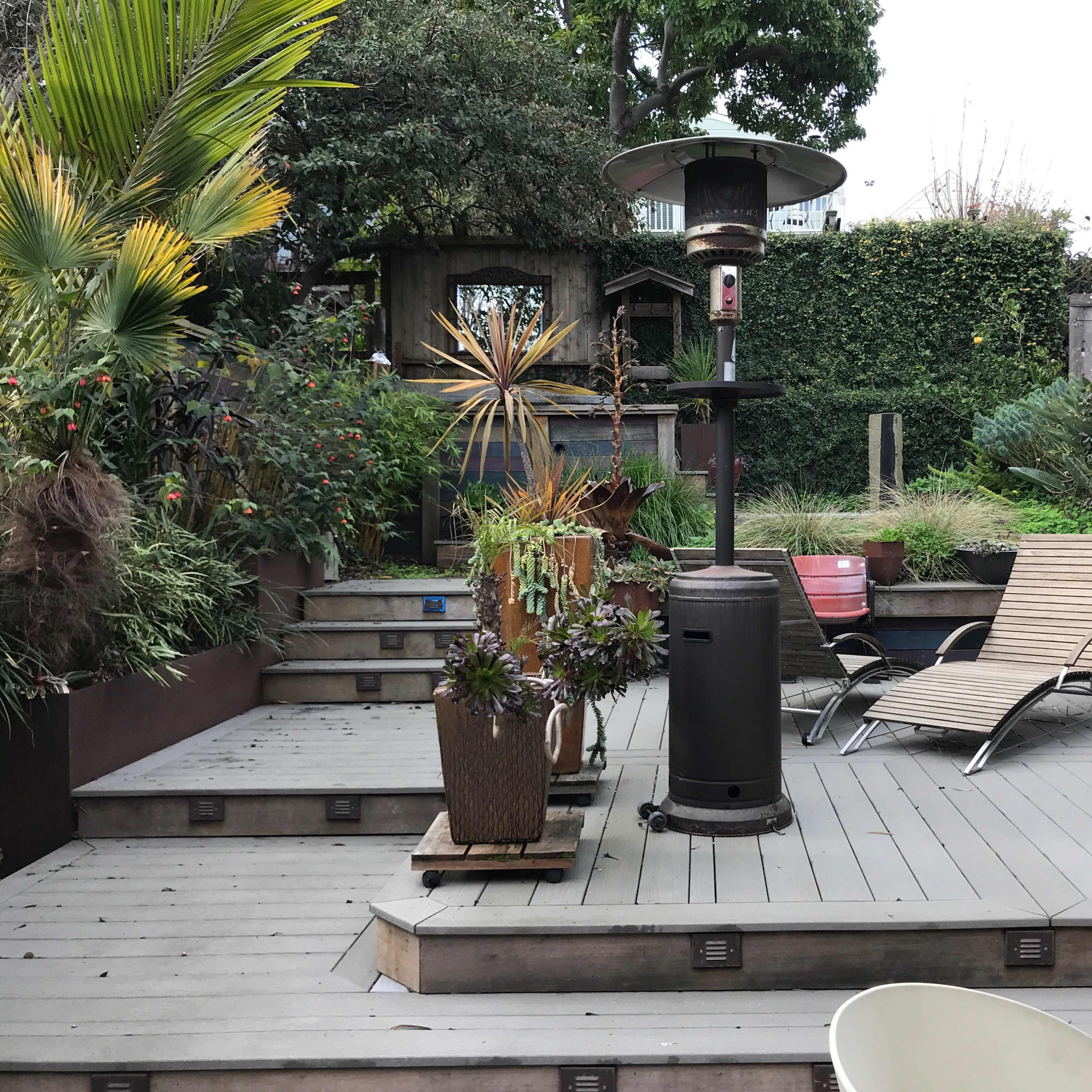 A landscaped patio area featuring a patio heater, lounge chairs, and various plants surrounding wooden decking and steps.